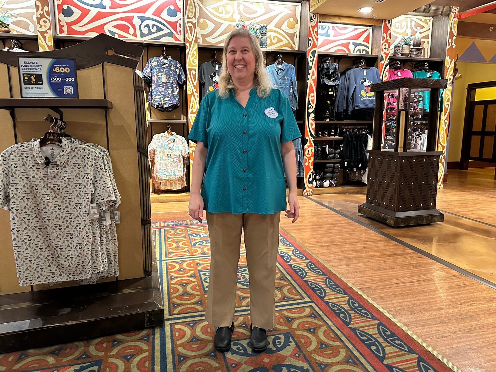 A cast member in a teal button-up shirt and khaki pants standing in a resort gift shop with racks of shirts and sweatshirts and colorful Polynesian-patterned wall panels behind