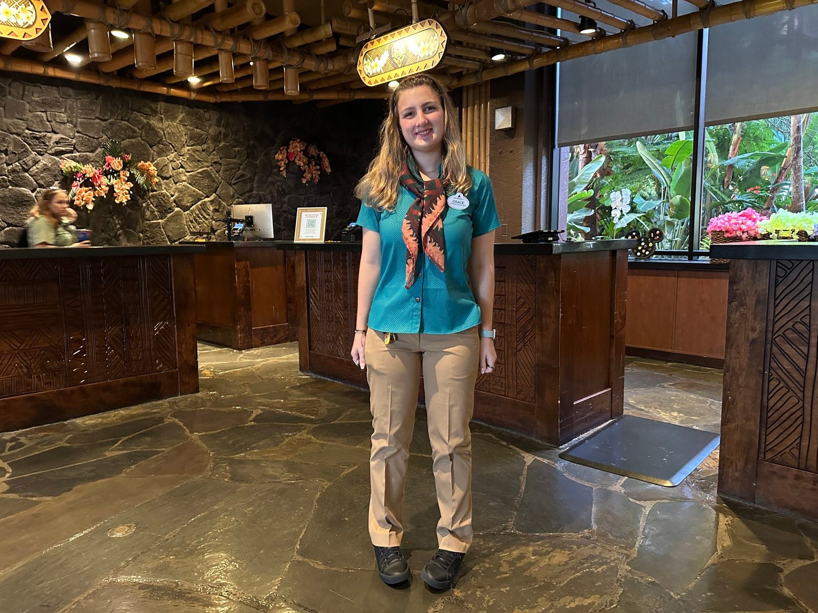 A front desk cast member in a teal short-sleeve top, an orange and green patterned scarf and khaki pants, standing in front of the carved wooden front desk in the Polynesian lobby