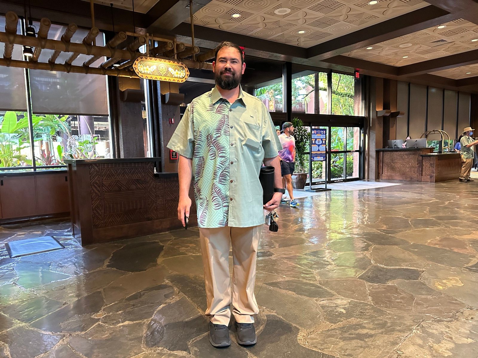 A smiling cast member standing in the Polynesian Village Resort lobby in a green button-up shirt with a leafy print on one half and khaki pants, with a carved wooden desk and stone floor behind him