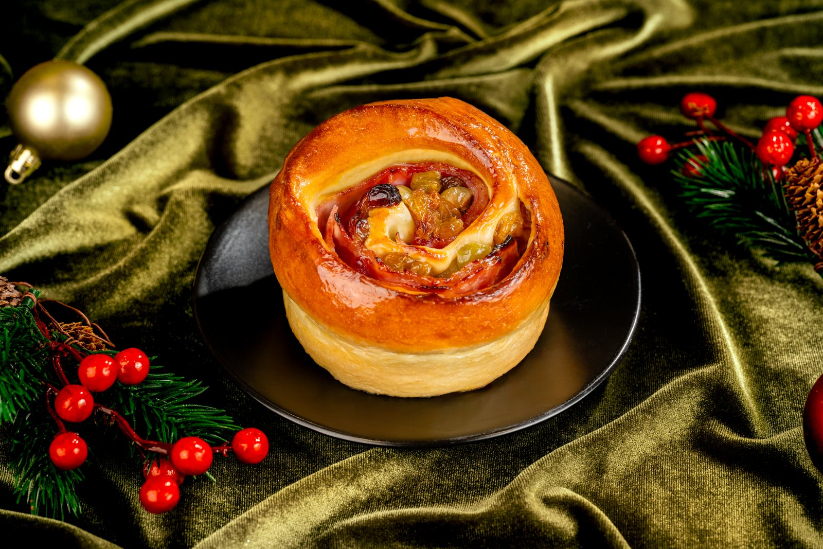 A round, glazed bread roll with a spiral of smoked ham, olives and raisins showing in the center, on a black plate