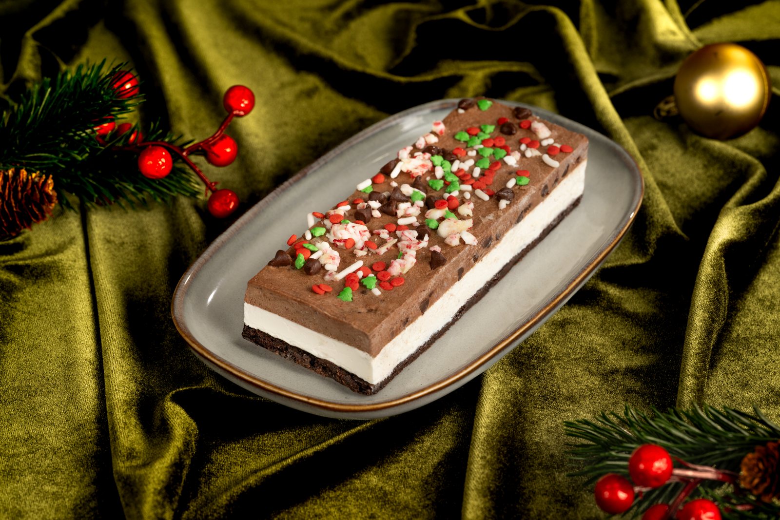 A rectangular ice cream sandwich with a layer of chocolate peppermint cookie dough on top, vanilla ice cream in the middle and brownie on the bottom, covered in red, white and green holiday sprinkles