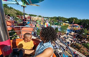 A child and an adult riding in an orange flower-pot seat high above Sesame Street Land, with colorful buildings, trees and crowds below