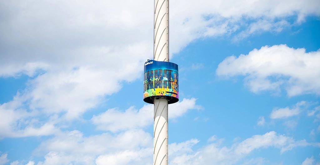 A tall white column against a blue sky with white clouds, with a round viewing cabin wrapped in a colorful graffiti-style mural partway up