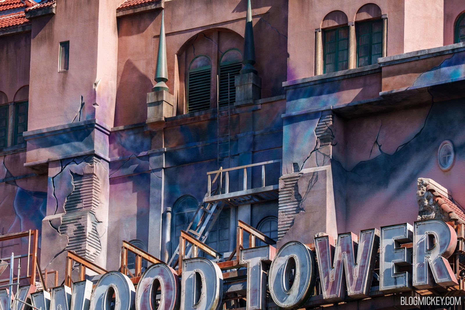 A close-up of the Hollywood Tower Hotel above its sign, with purple and blue airbrushed streaks, painted cracks and broken stonework on pink stucco, a metal ladder leaning to a small balcony, and the large WOOD TOWER letters of the sign below
