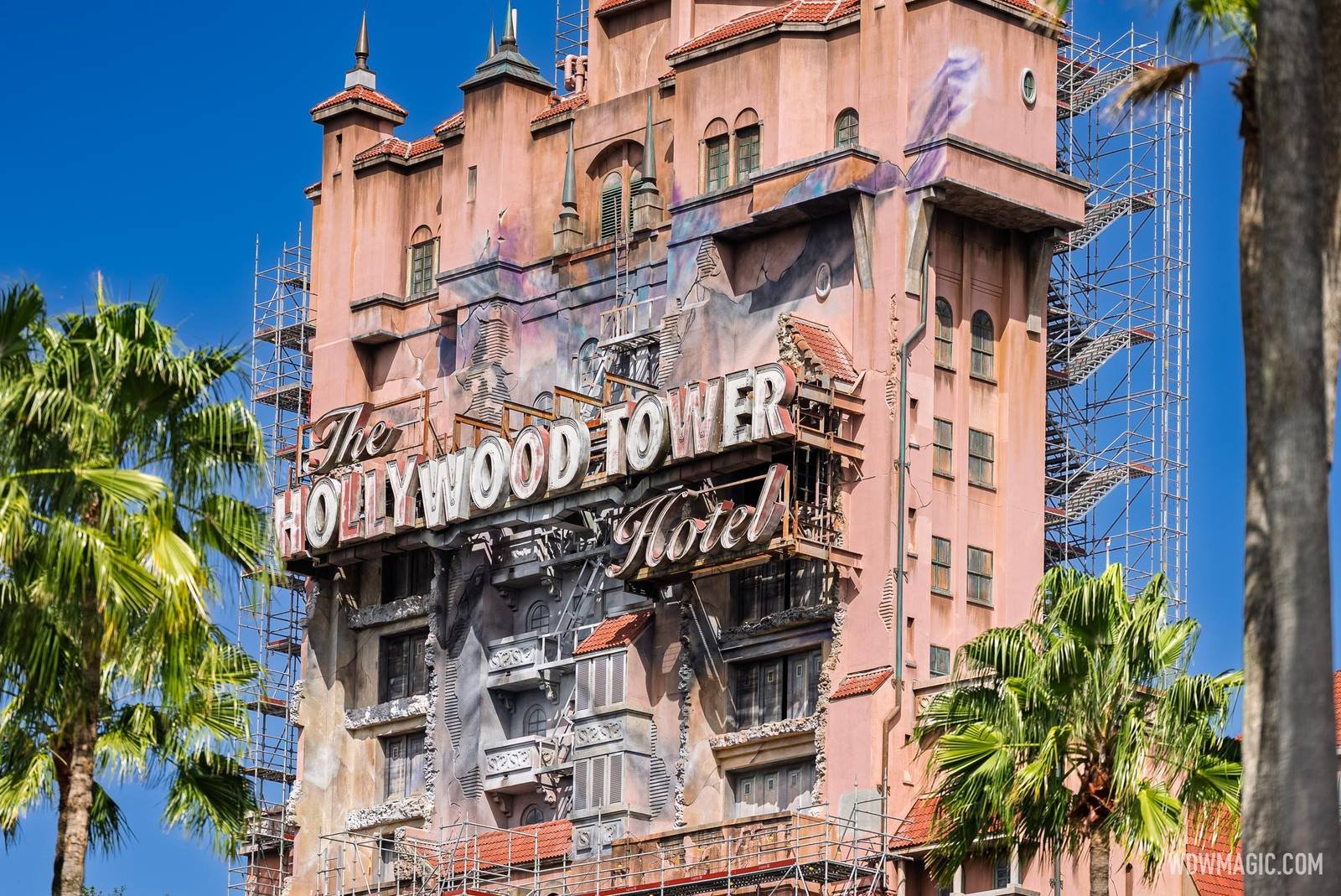 The upper front of the pink Hollywood Tower Hotel with purple and blue streaks and painted cracks spreading above and around The Hollywood Tower Hotel sign, with scaffolding still standing at both edges and across the lower floors, and palm trees in the foreground