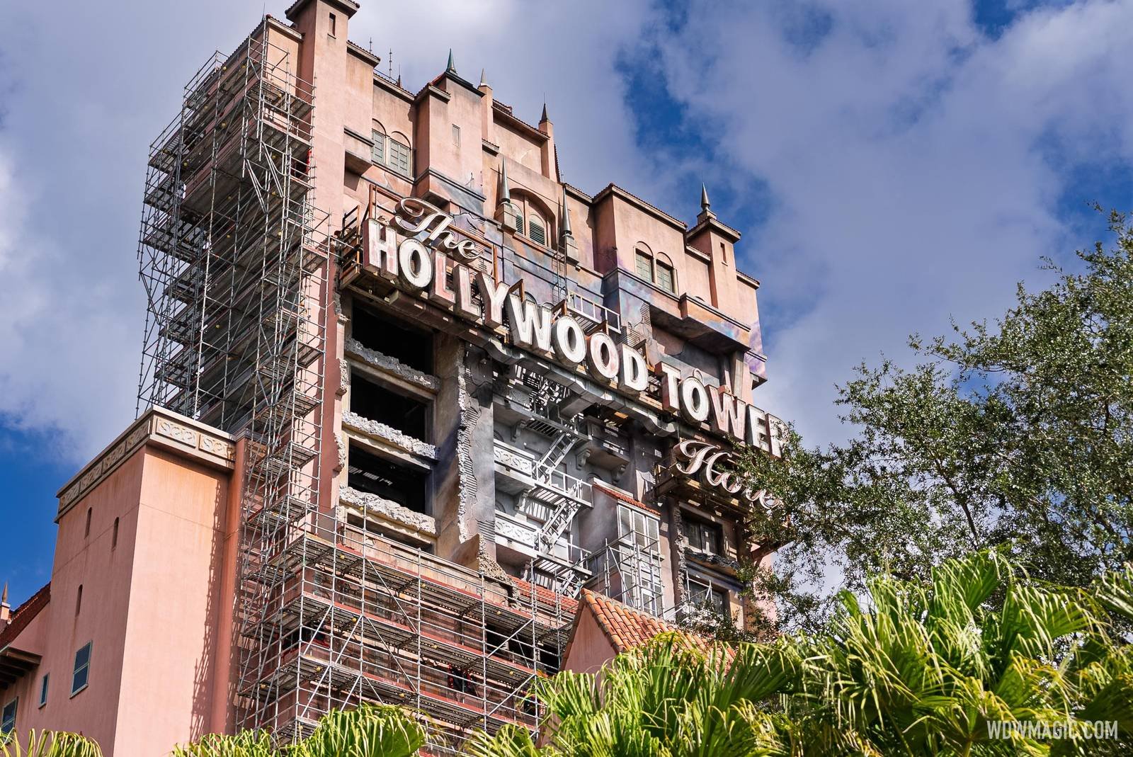 Looking up at the Hollywood Tower Hotel from below, with a tall stack of scaffolding running up the left side of the building beside the repainted facade and The Hollywood Tower Hotel sign, under a partly cloudy sky