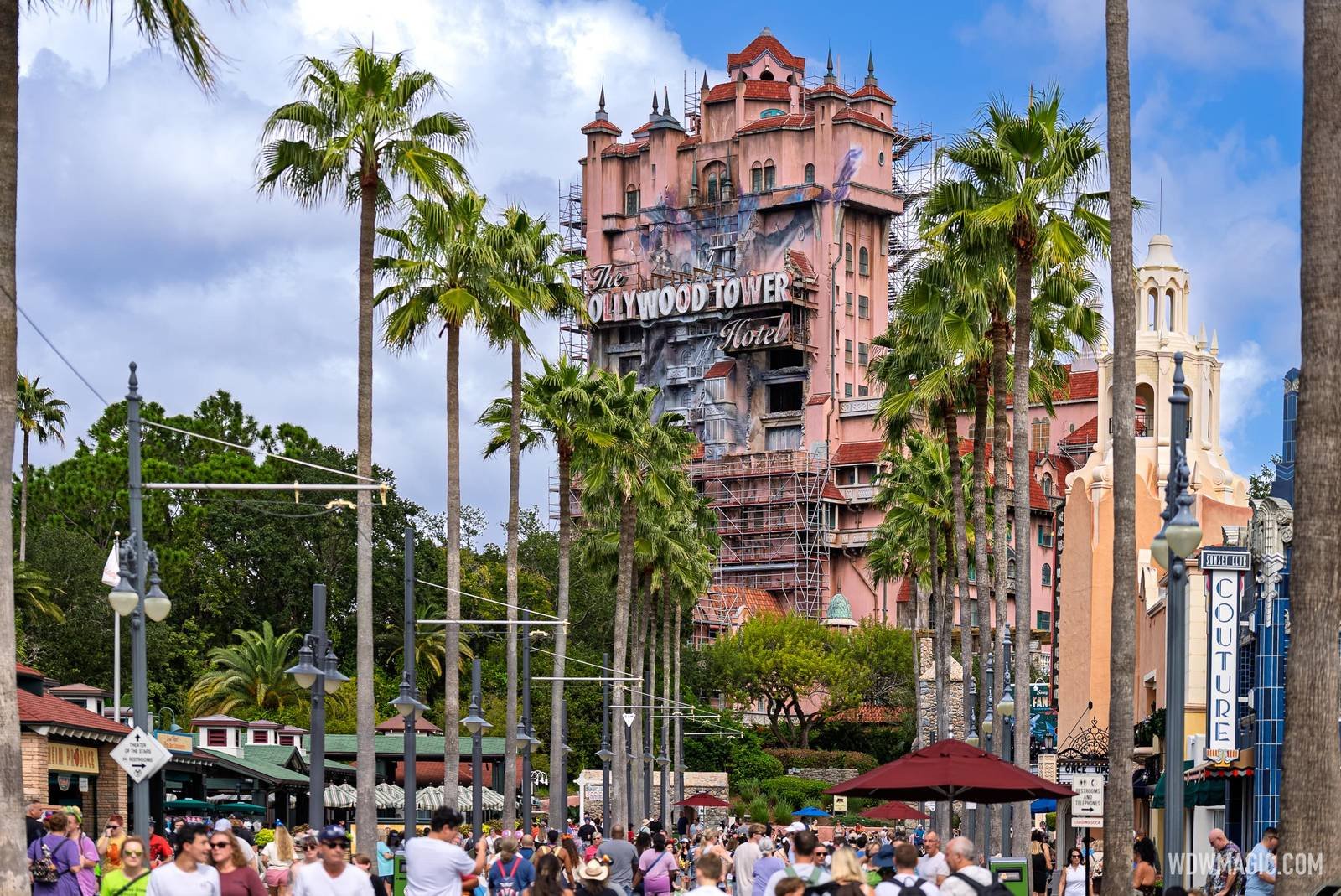 A crowd of guests on Sunset Boulevard below the Hollywood Tower Hotel, which rises above the palm trees with its sign and repainted upper floors visible and scaffolding on its lower levels and up both sides