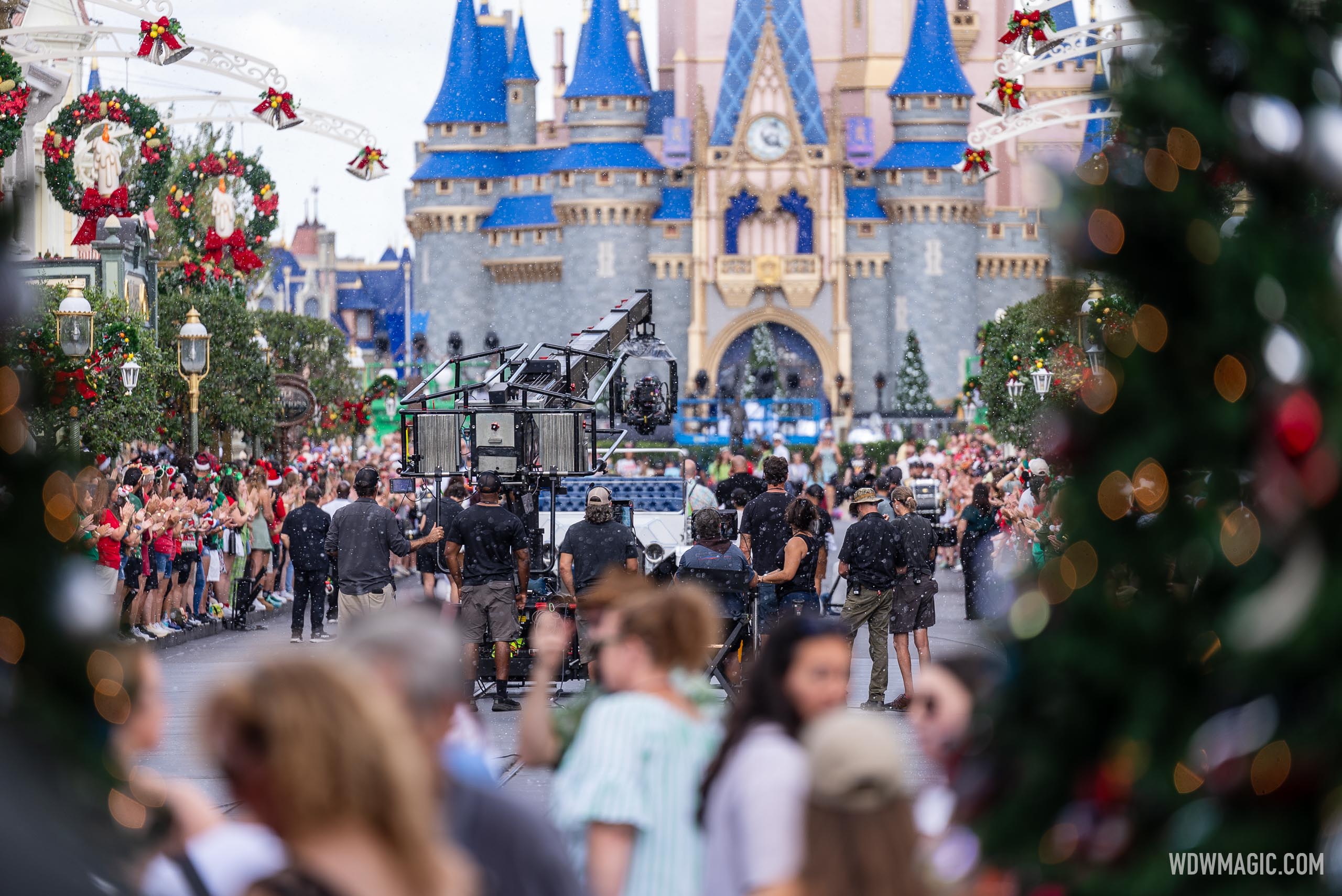 A film crew operates a large camera crane in the middle of a decorated Main Street, U.S.A. with Cinderella Castle behind it and a crowd in holiday outfits lining both sides of the street