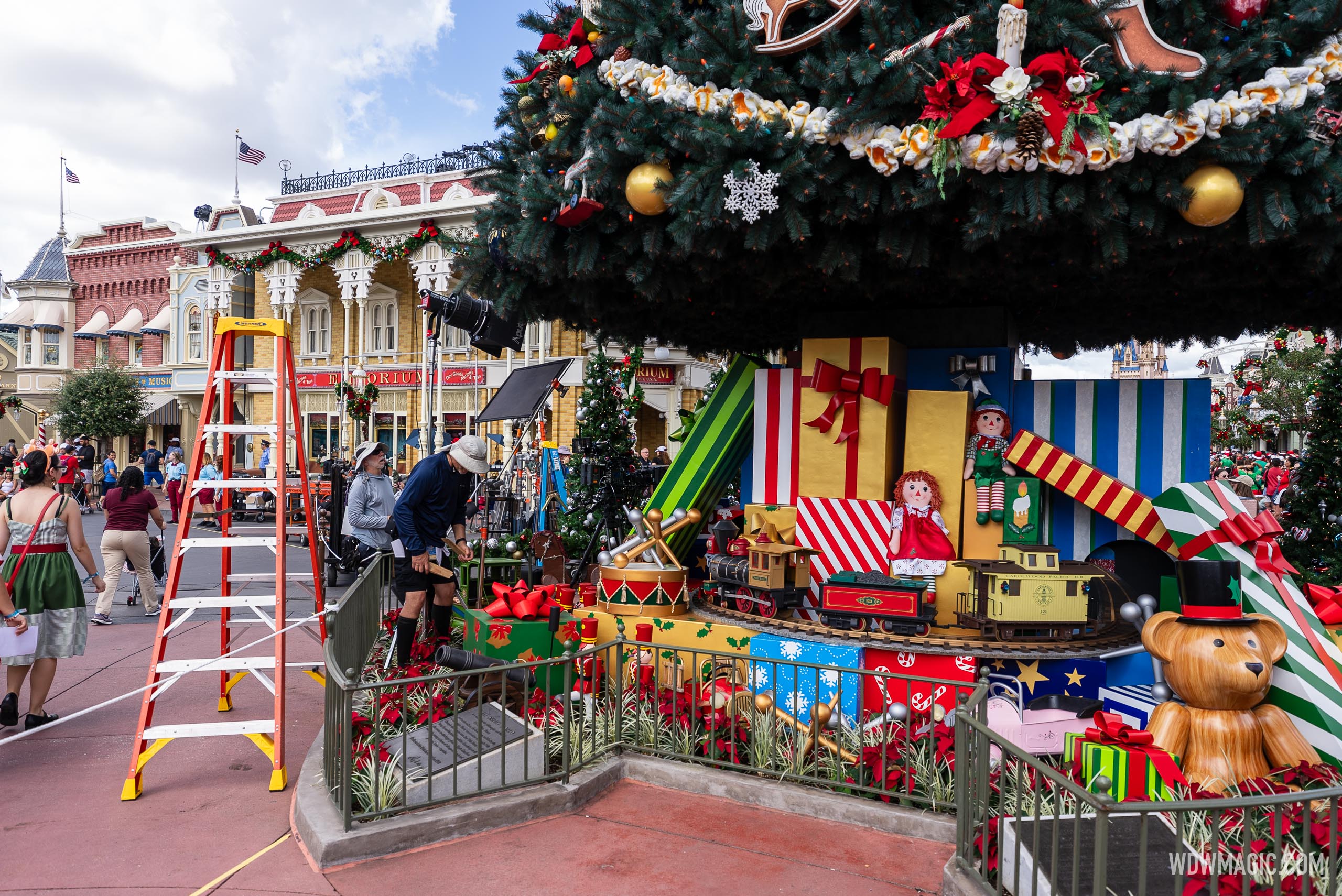Crew members set up lights and equipment beside the decorated Town Square Christmas tree at Magic Kingdom, with an orange ladder in the foreground and the Emporium behind