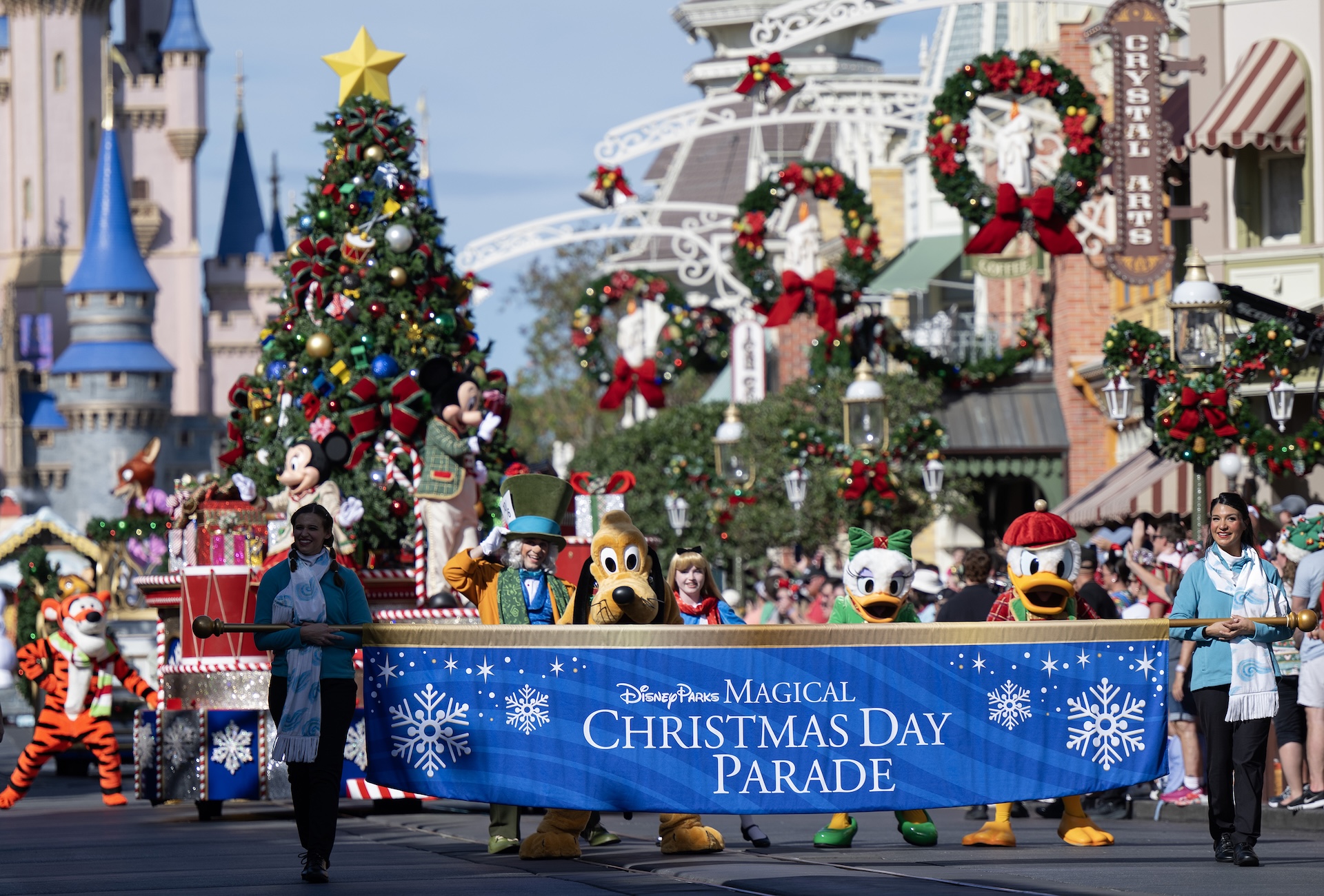 Performers carry a blue Disney Parks Magical Christmas Day Parade banner down Main Street, U.S.A., followed by Pluto, Daisy Duck, Donald Duck, the Mad Hatter, Alice and Tigger, with Mickey and Minnie on a Christmas tree float