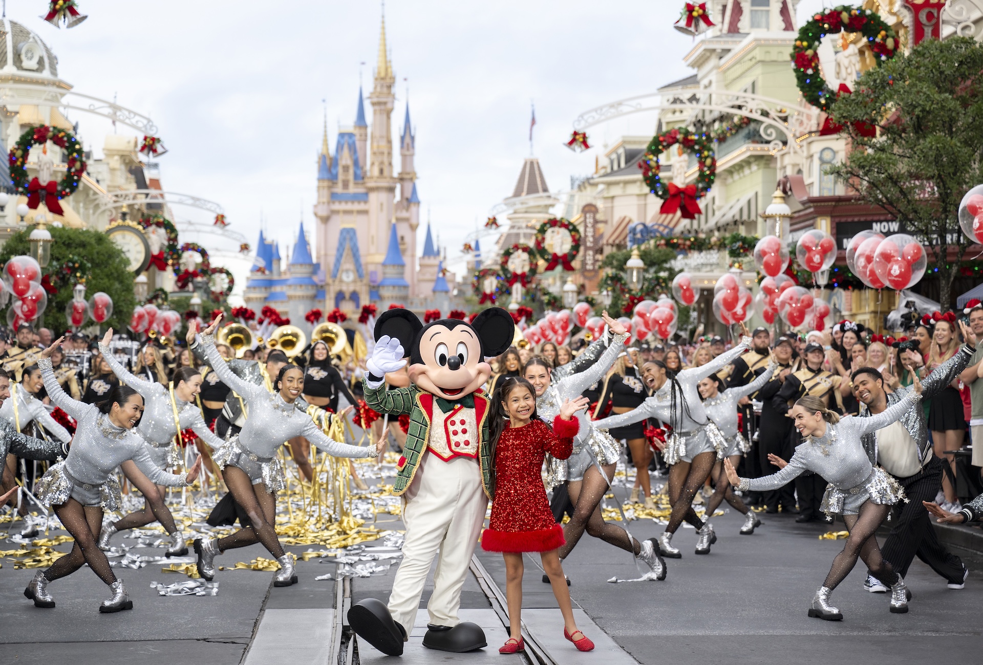 Mickey Mouse in a green plaid holiday jacket waves on Main Street, U.S.A. beside a girl in a red sequined dress, surrounded by dancers in silver costumes with Cinderella Castle behind them