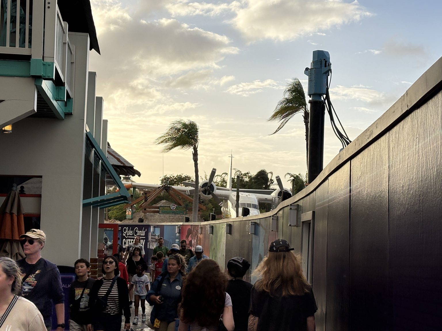 A crowd of guests walks a narrow path between a building and a tall dark construction wall, with the Lone Palm Airport seaplane and palm trees ahead