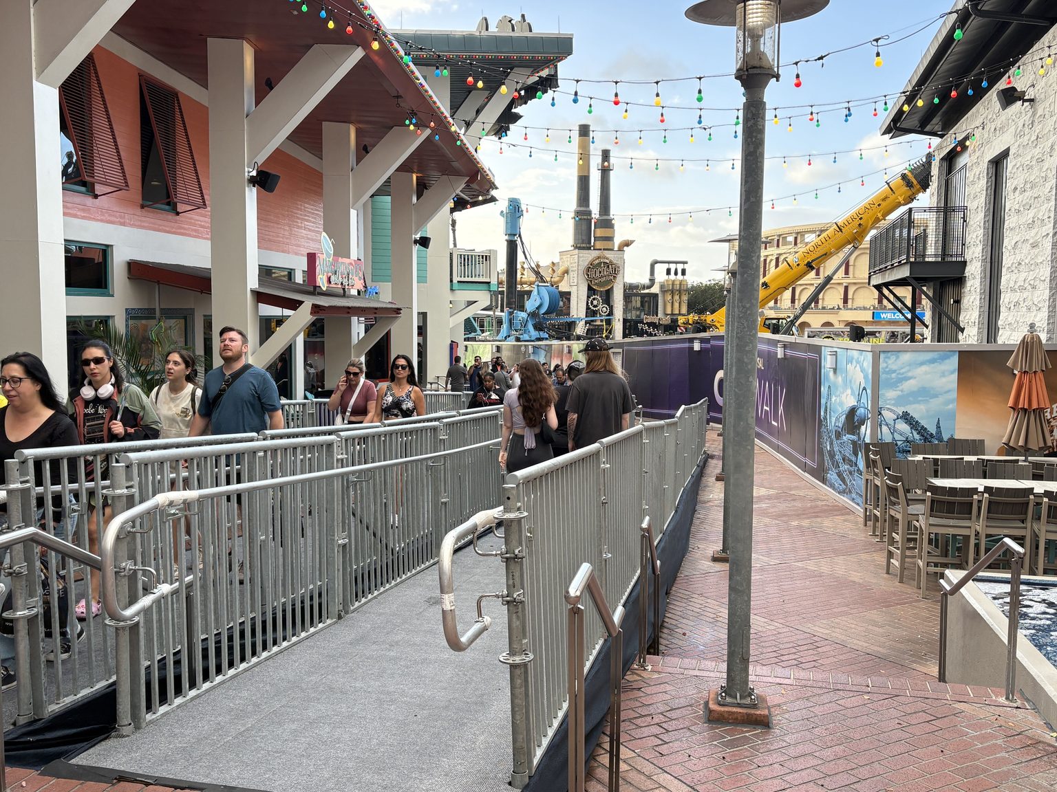 Guests walk along a gray temporary ramp with metal railings beside a brick walkway, with construction walls and a yellow crane on the right