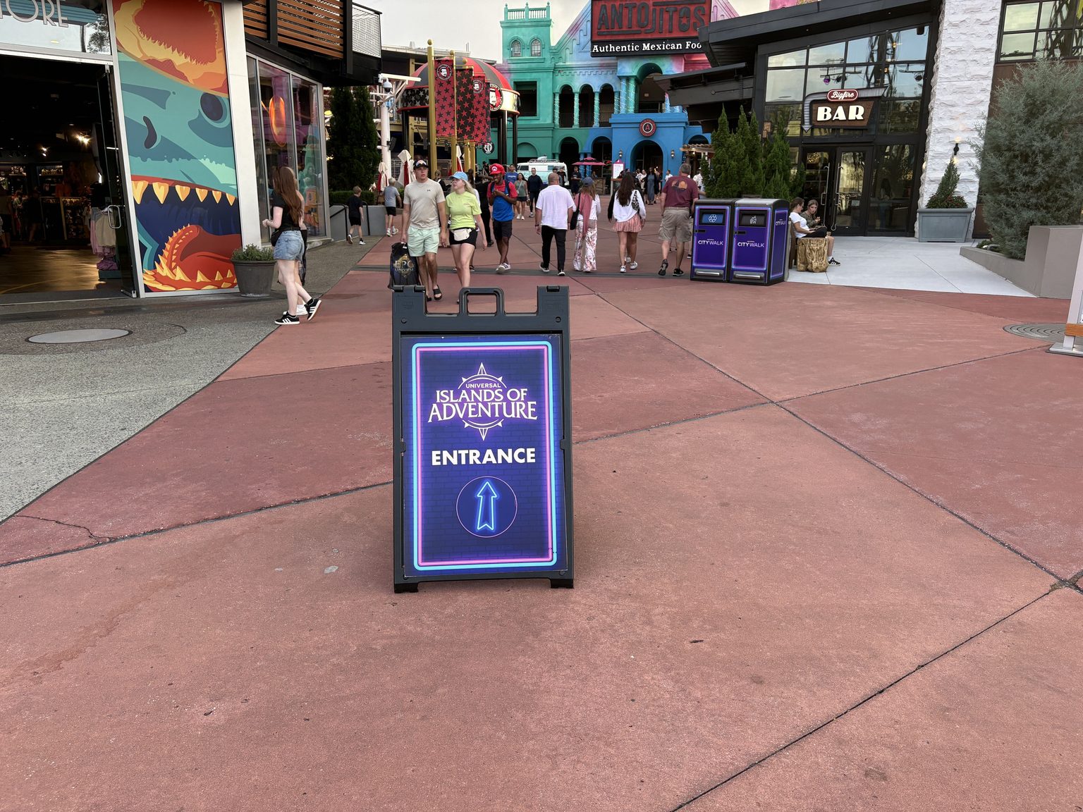 A purple Universal Islands of Adventure Entrance sandwich-board sign with an upward arrow on the CityWalk pavement, with the Antojitos building and a Bigfire bar sign in the background