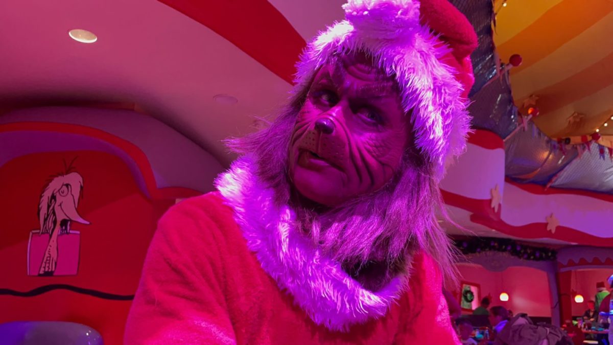 Close-up of the Grinch in a fuzzy Santa hat and red suit leaning toward the camera inside a brightly colored dining room with red and white striped walls and a Seuss character mural