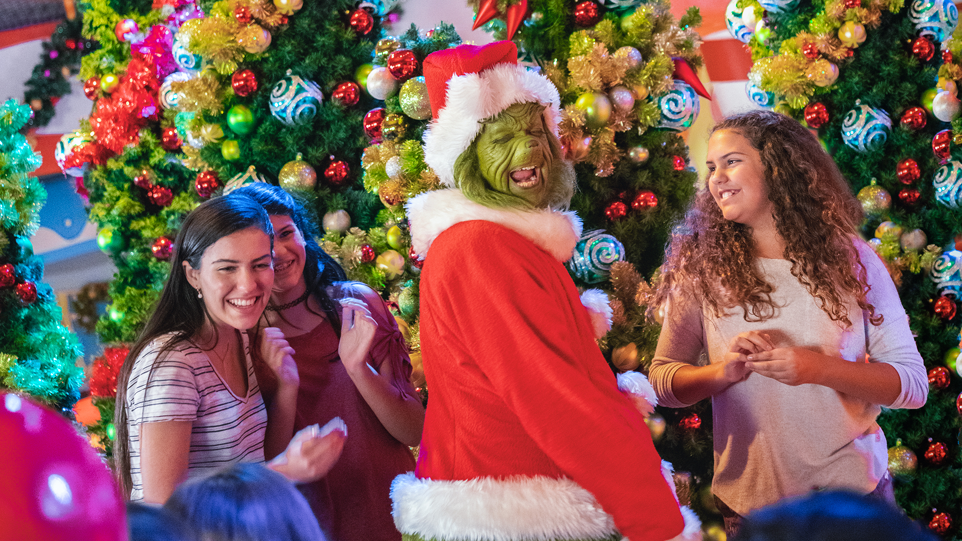 The Grinch in a red Santa suit and hat, laughing with three smiling guests in front of Christmas trees covered in red, gold and teal ornaments
