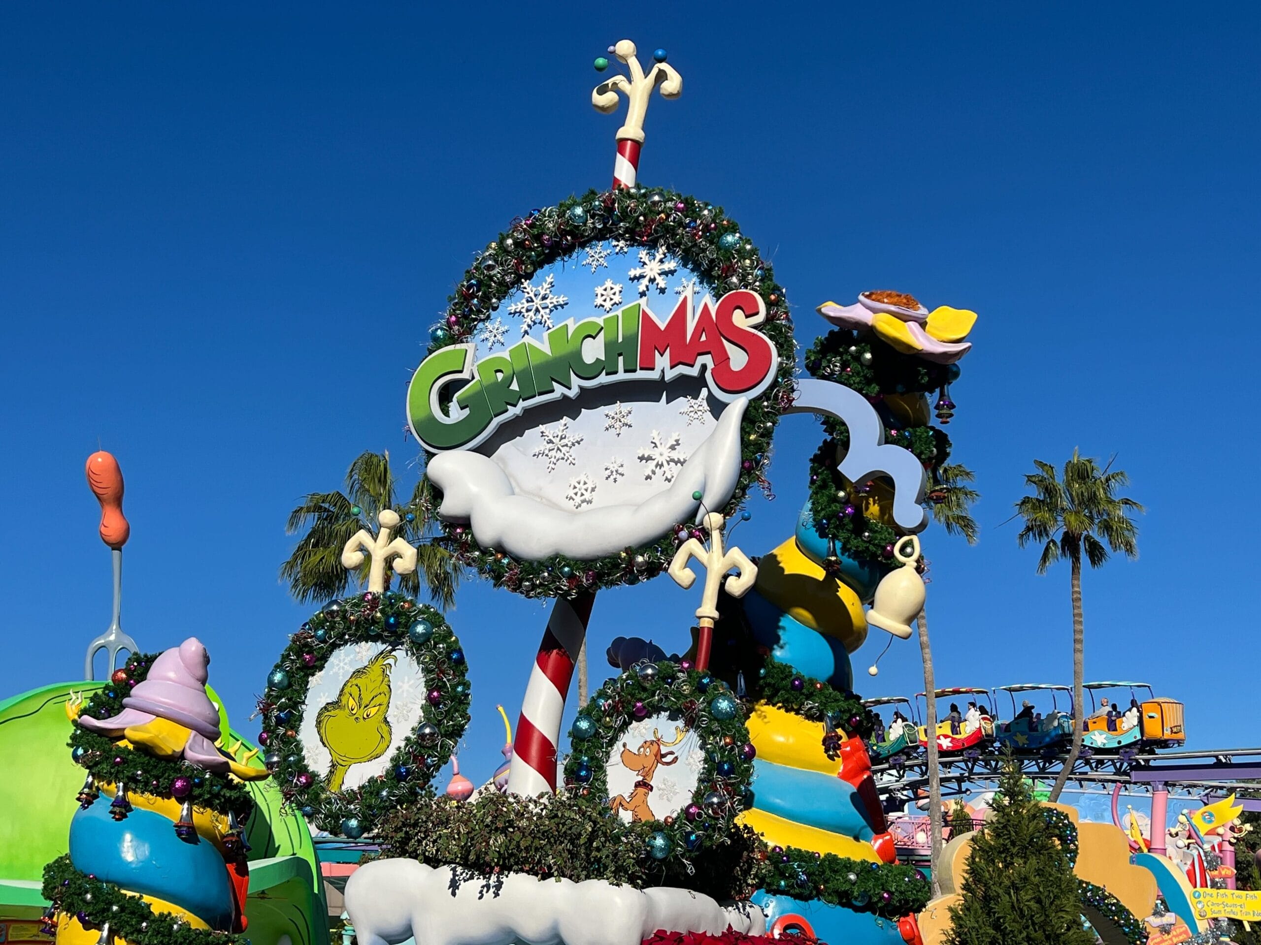 A large round Grinchmas sign wrapped in garland with snowflakes, flanked by smaller wreaths showing the Grinch and his dog Max, with palm trees, a blue sky and a Seuss Landing trolley ride in the background