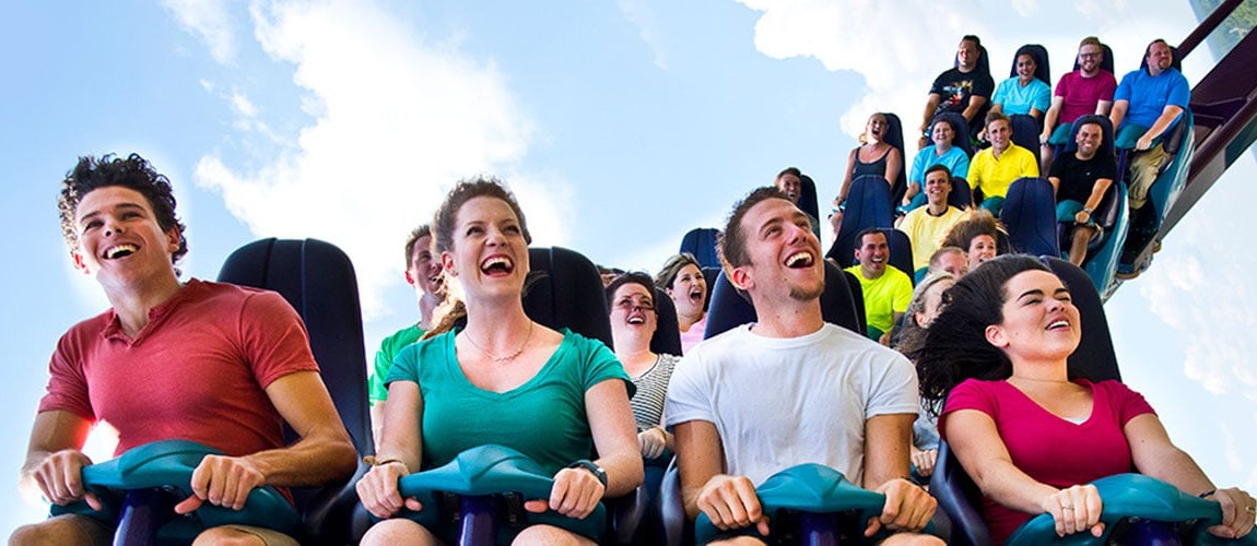 Smiling and screaming riders in the front rows of the Mako coaster train, with the train stretching up the track behind them against a blue sky
