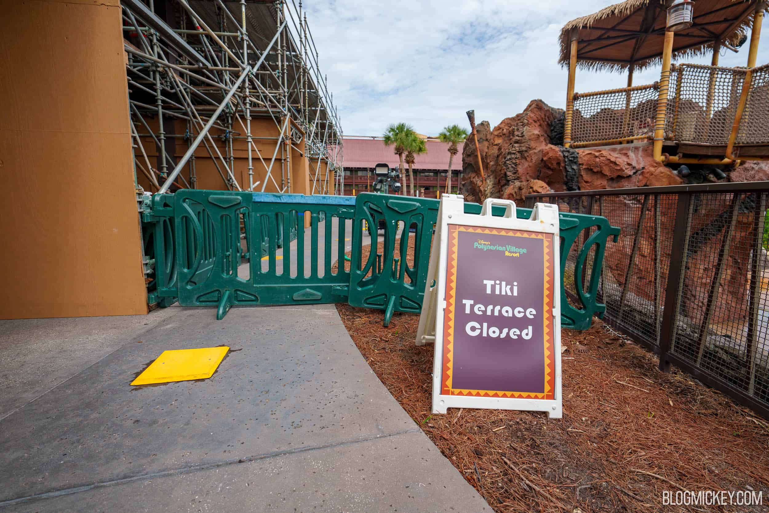 A purple sandwich-board sign reading Tiki Terrace Closed with the Disney's Polynesian Village Resort logo, standing in front of green plastic barricades, with scaffolding along a building wall at left and rockwork at right