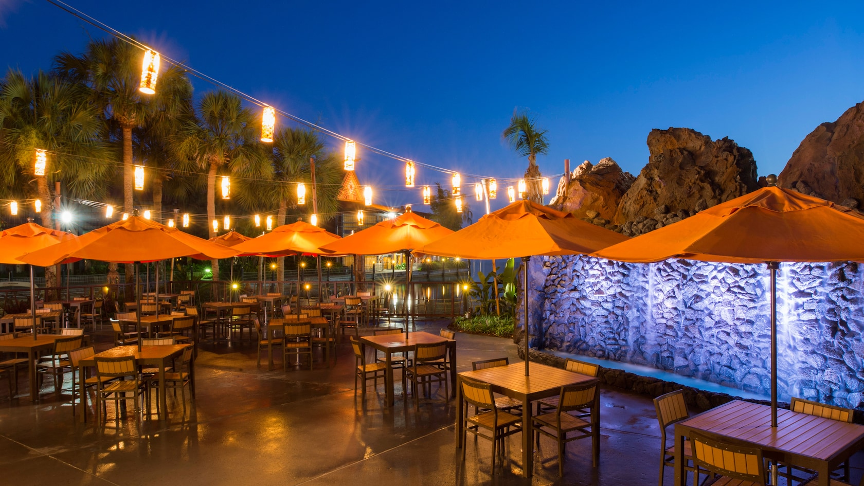 Trader Sam's Tiki Terrace at night, with orange umbrellas over empty tables, strings of glowing lanterns overhead, palm trees, and a lit rock wall with a waterfall at right