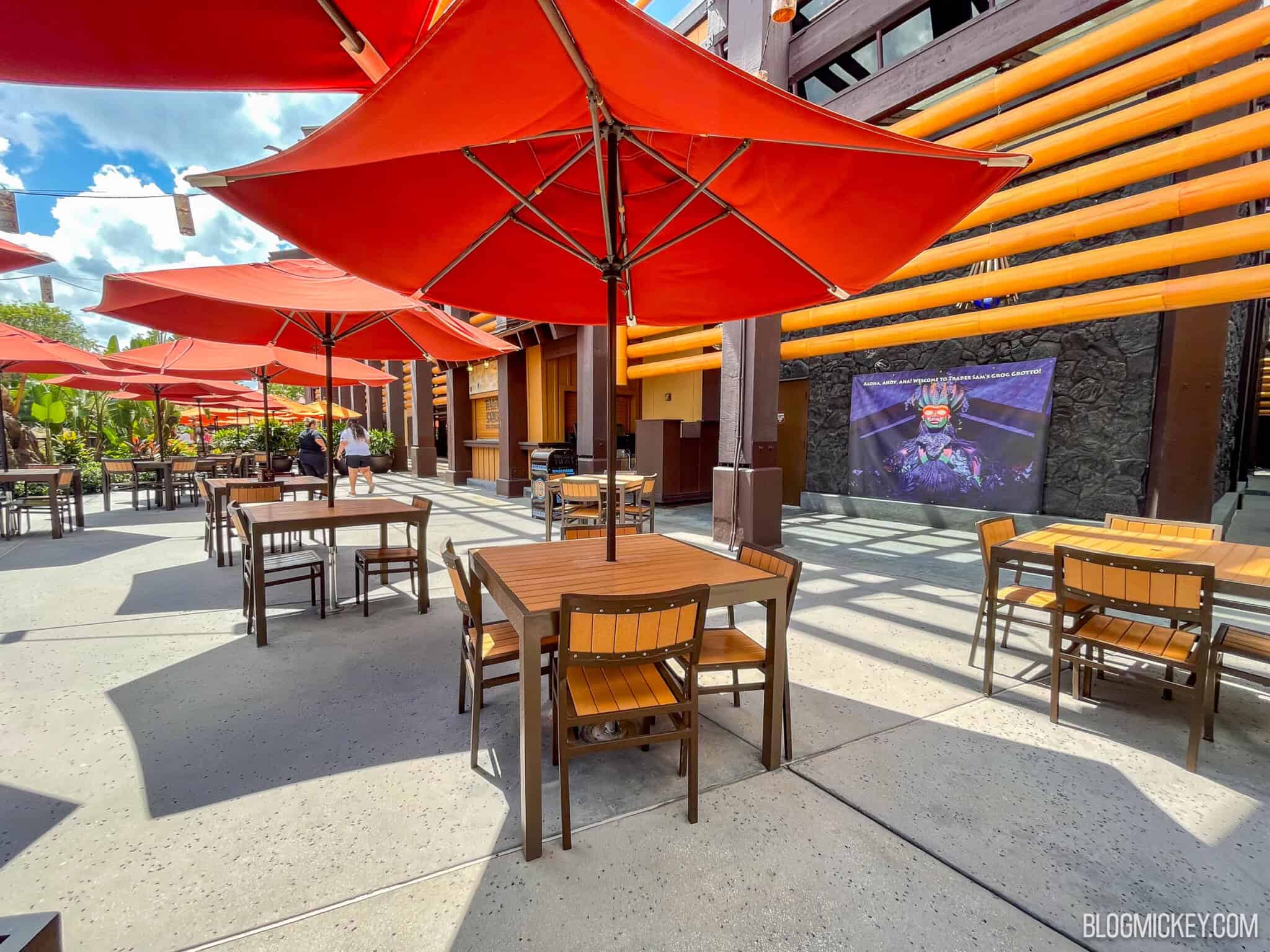 Wooden patio tables and chairs under red-orange umbrellas on a sunny day, beside a building with orange bamboo-style beams and a banner welcoming guests to Trader Sam's Grog Grotto
