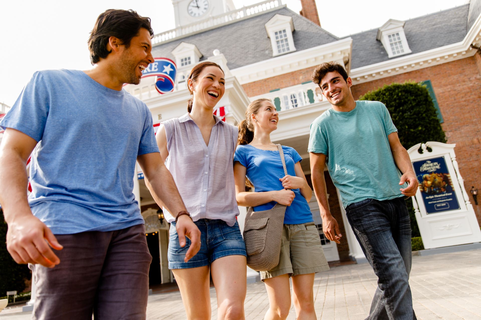 Four smiling friends walk in front of the red brick American Adventure building at EPCOT, with its white clock tower and an American Adventure show poster at right