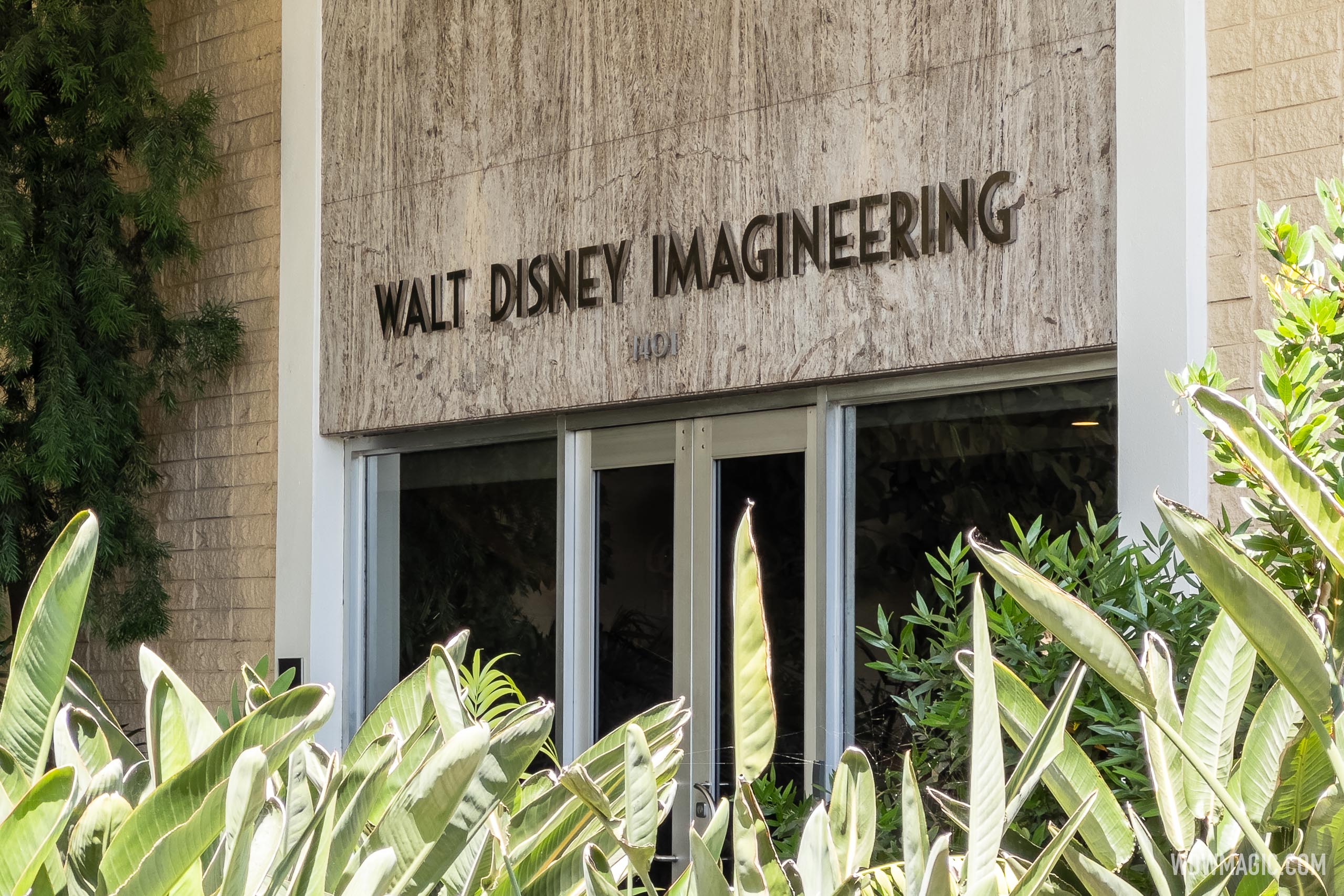 The Walt Disney Imagineering name in metal letters on a marble panel above glass entrance doors, with the street number 1401 below it and broad green leaves in the foreground