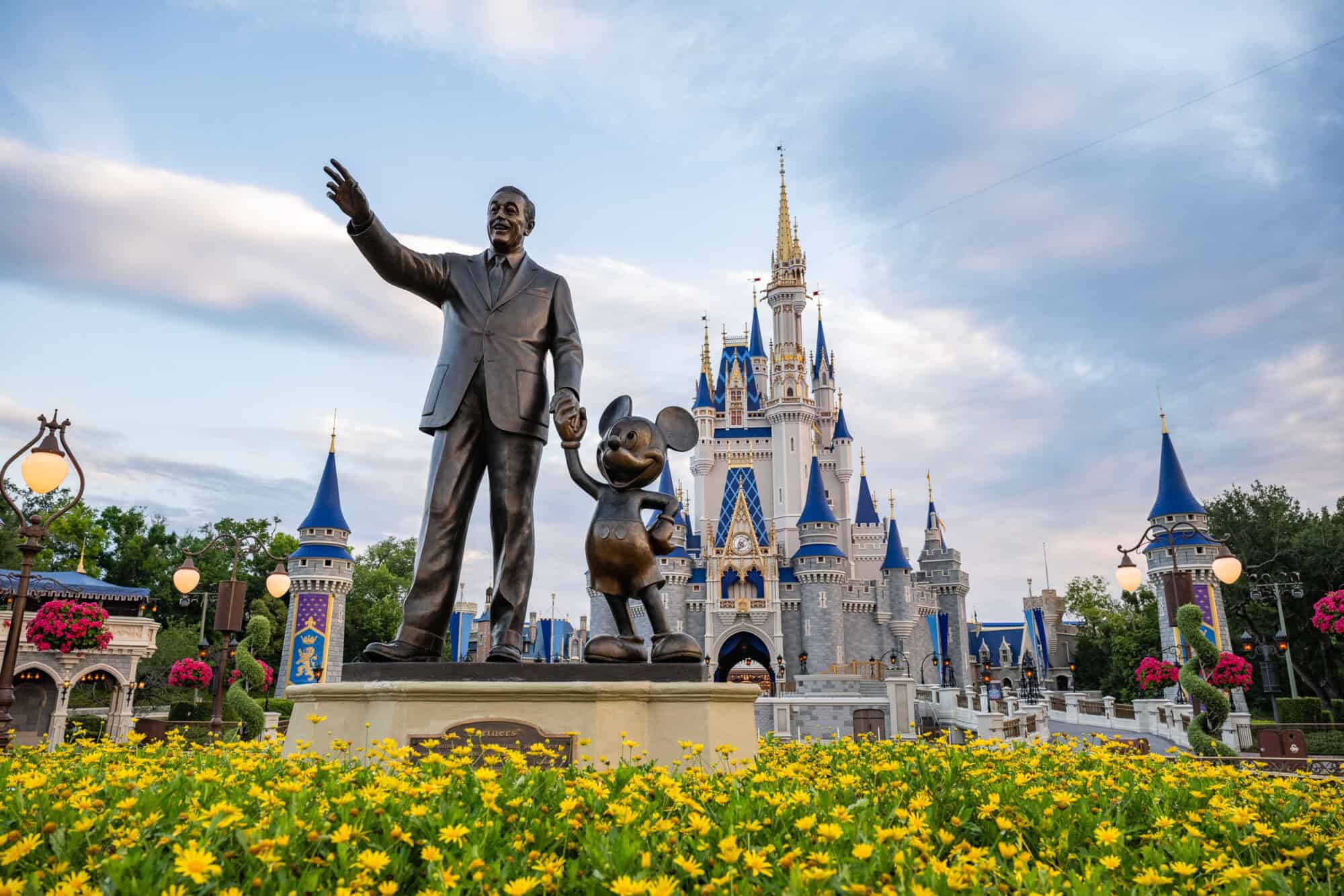 The bronze Partners statue of Walt Disney holding Mickey Mouse's hand in front of Cinderella Castle, with yellow flowers in the foreground