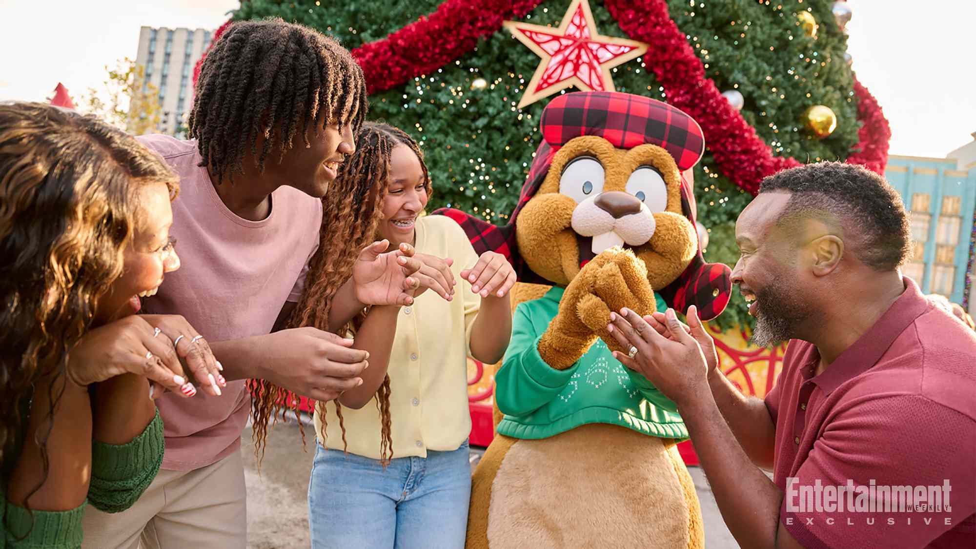 Earl the Squirrel, a costumed character in a red plaid hat with ear flaps and a green sweater, laughs with a family of four in front of a large Christmas tree topped with a red and gold star