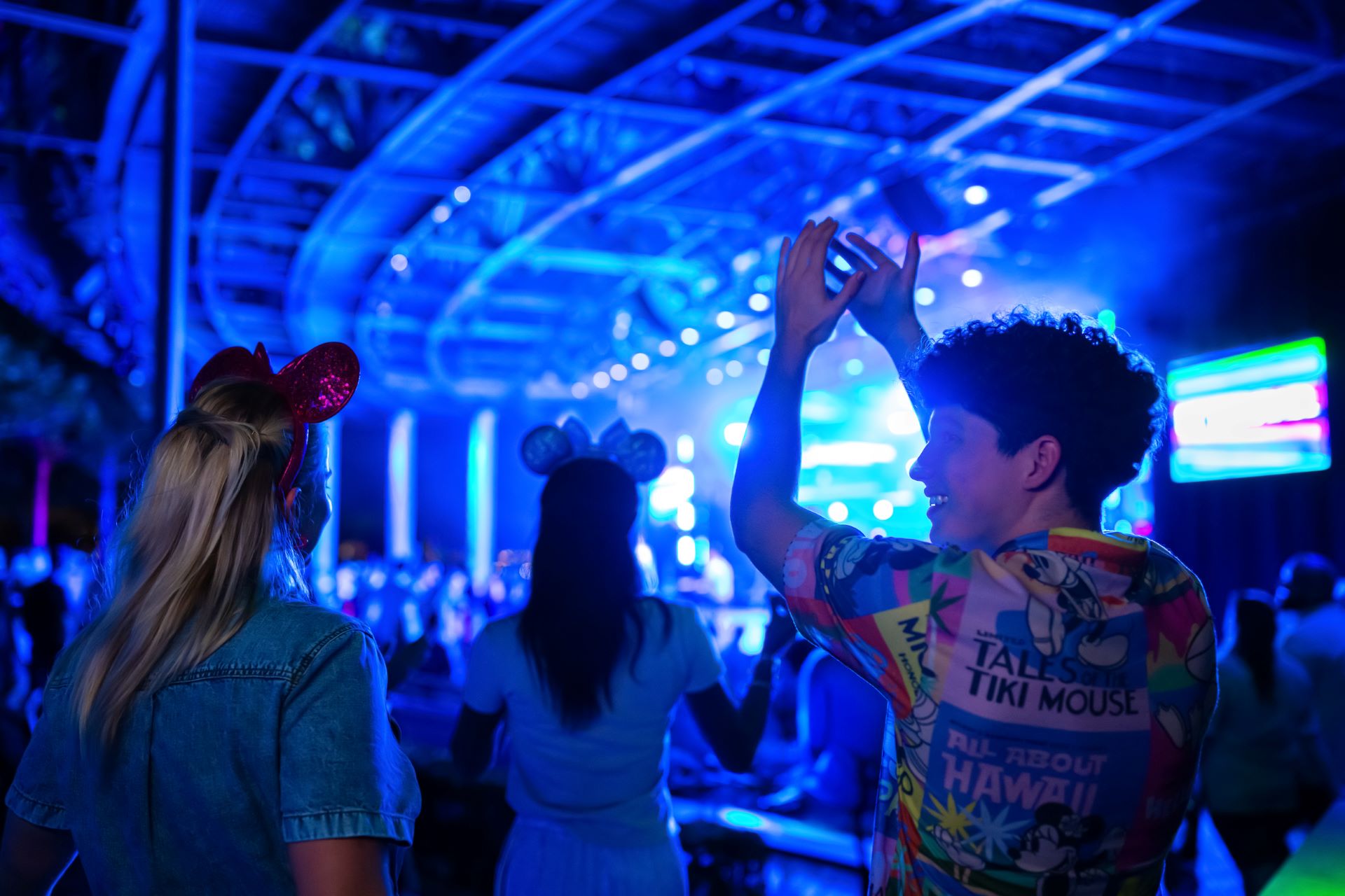 A guest raises his hands toward a stage lit in deep blue under a curved metal canopy at night, beside two guests in Minnie ears