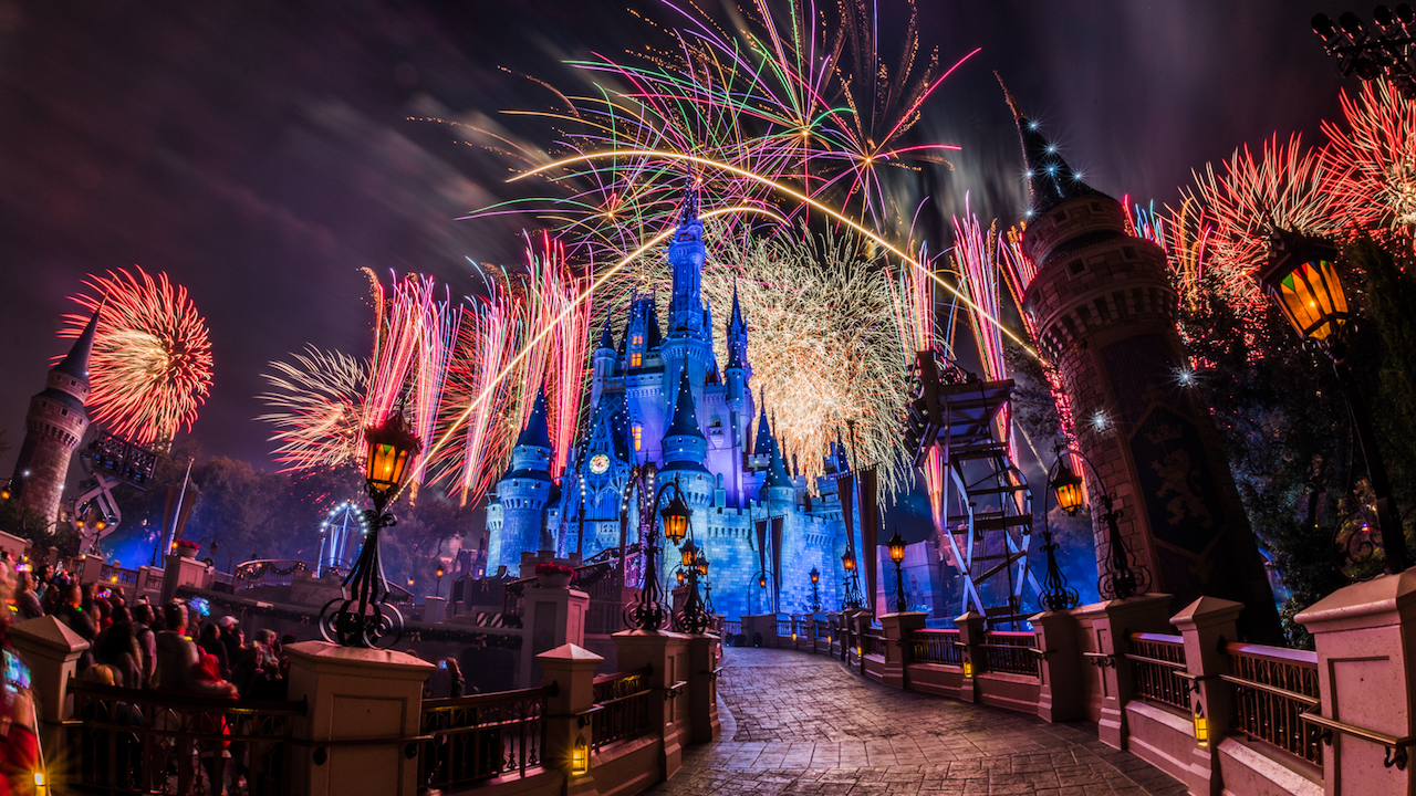 Pink, gold and white fireworks explode over a blue-lit Cinderella Castle, seen from the curved castle walkway lined with lampposts, with guests watching at left