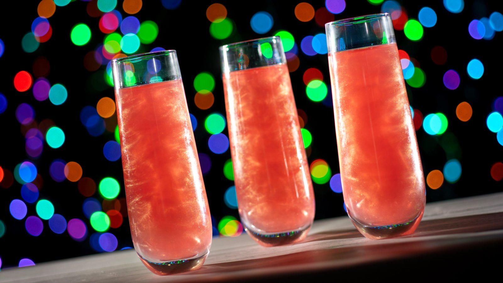 Three tall stemless flutes filled with a sparkling pink drink swirled with glitter, set against colorful blurred holiday lights