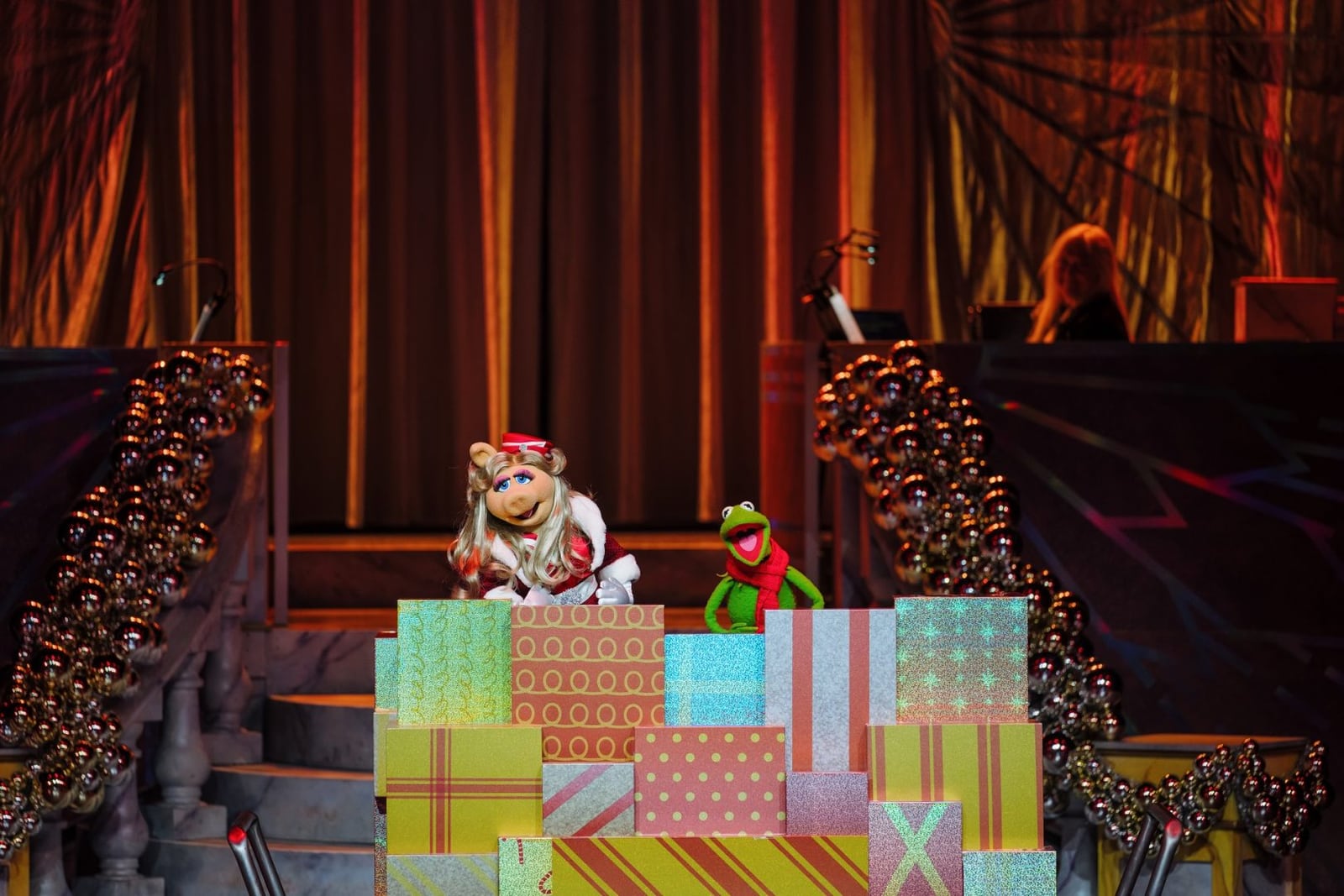 Miss Piggy in a Santa hat and Kermit the Frog in a red scarf on top of a stack of wrapped gift boxes on a stage framed by gold curtains and ornament garlands