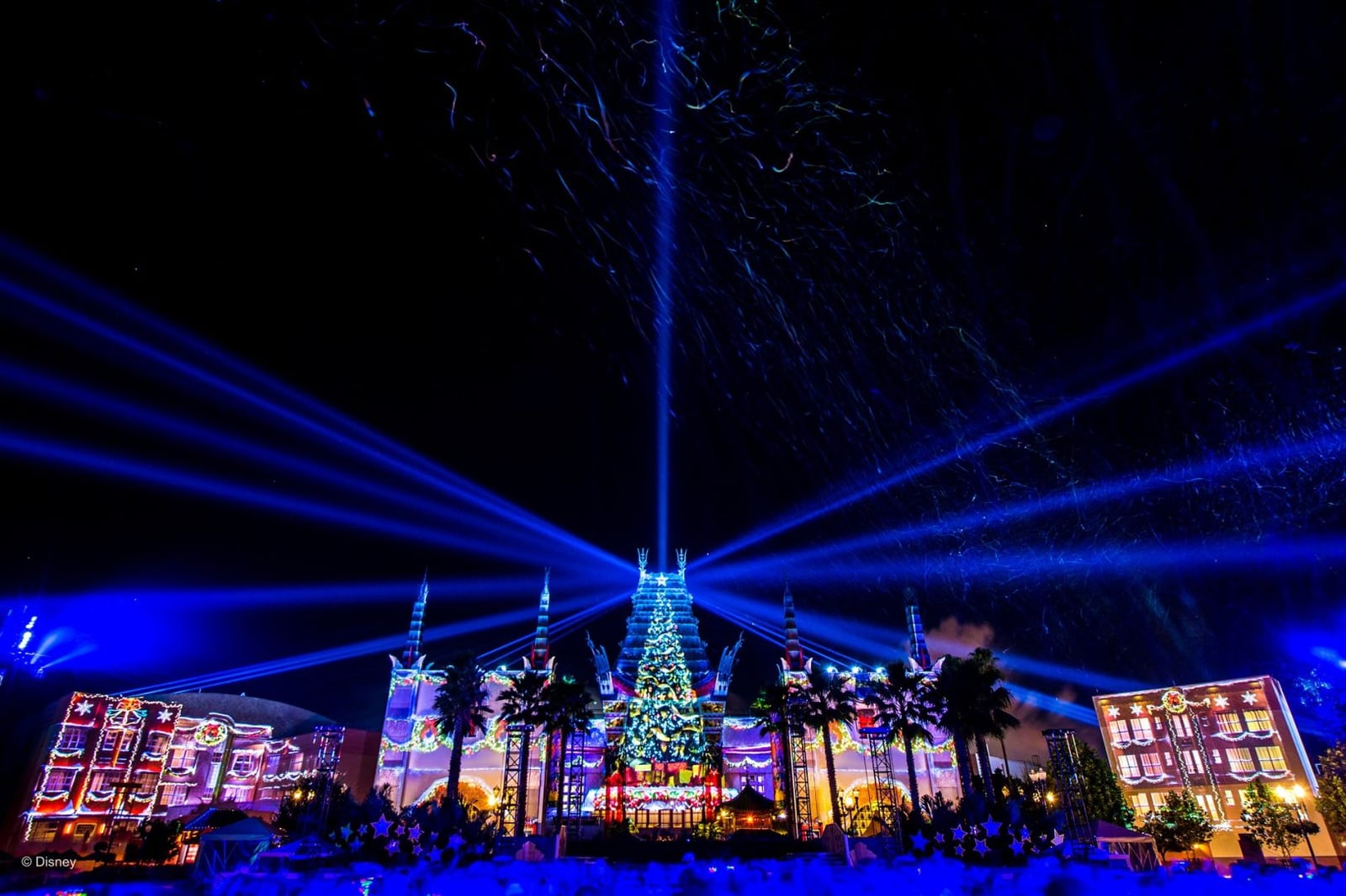 Blue lasers fan out from the Chinese Theater at Hollywood Studios at night, with a lit Christmas tree projected on its front, holiday projections on the buildings on either side and snow effects in the air