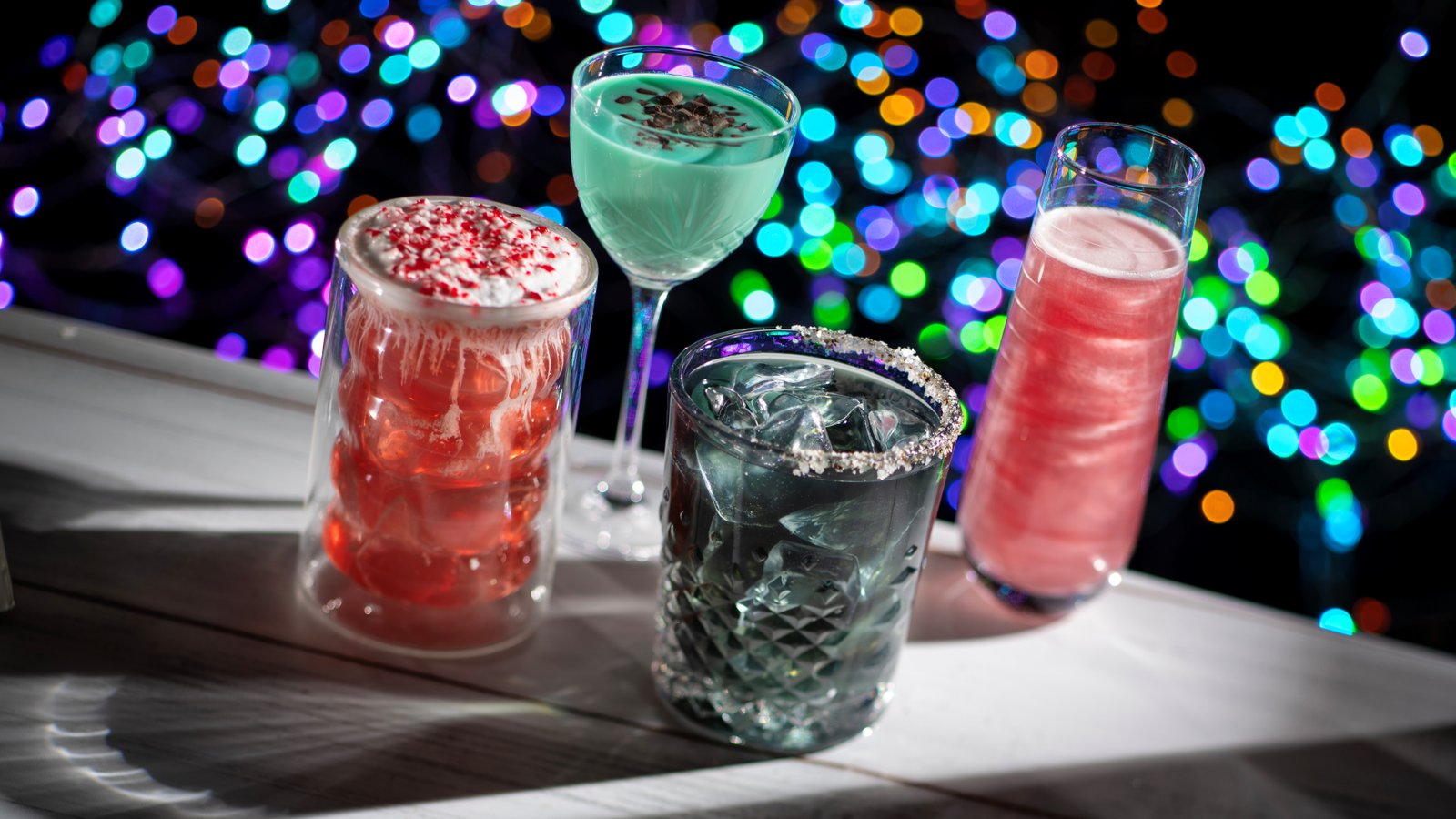 Four holiday cocktails on a white bar against a backdrop of colorful blurred lights: a red layered drink topped with crushed peppermint, a mint green drink in a coupe, a dark drink on the rocks with a salted rim and a sparkling pink drink in a tall glass