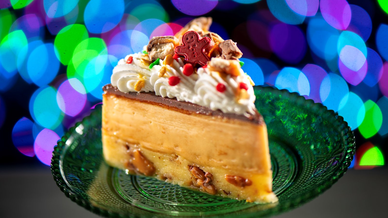 A slice of pie with a caramel layer, chocolate ganache and whipped cream, topped with candy pieces, holiday sprinkles and a red gingerbread man, on a green glass plate