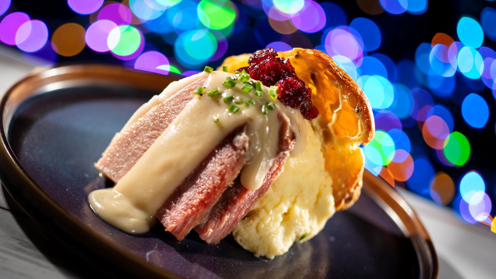 Slices of ham with gravy, mashed potatoes and a spoonful of cranberry chutney on a golden popover, served on a dark plate in front of blurred holiday lights