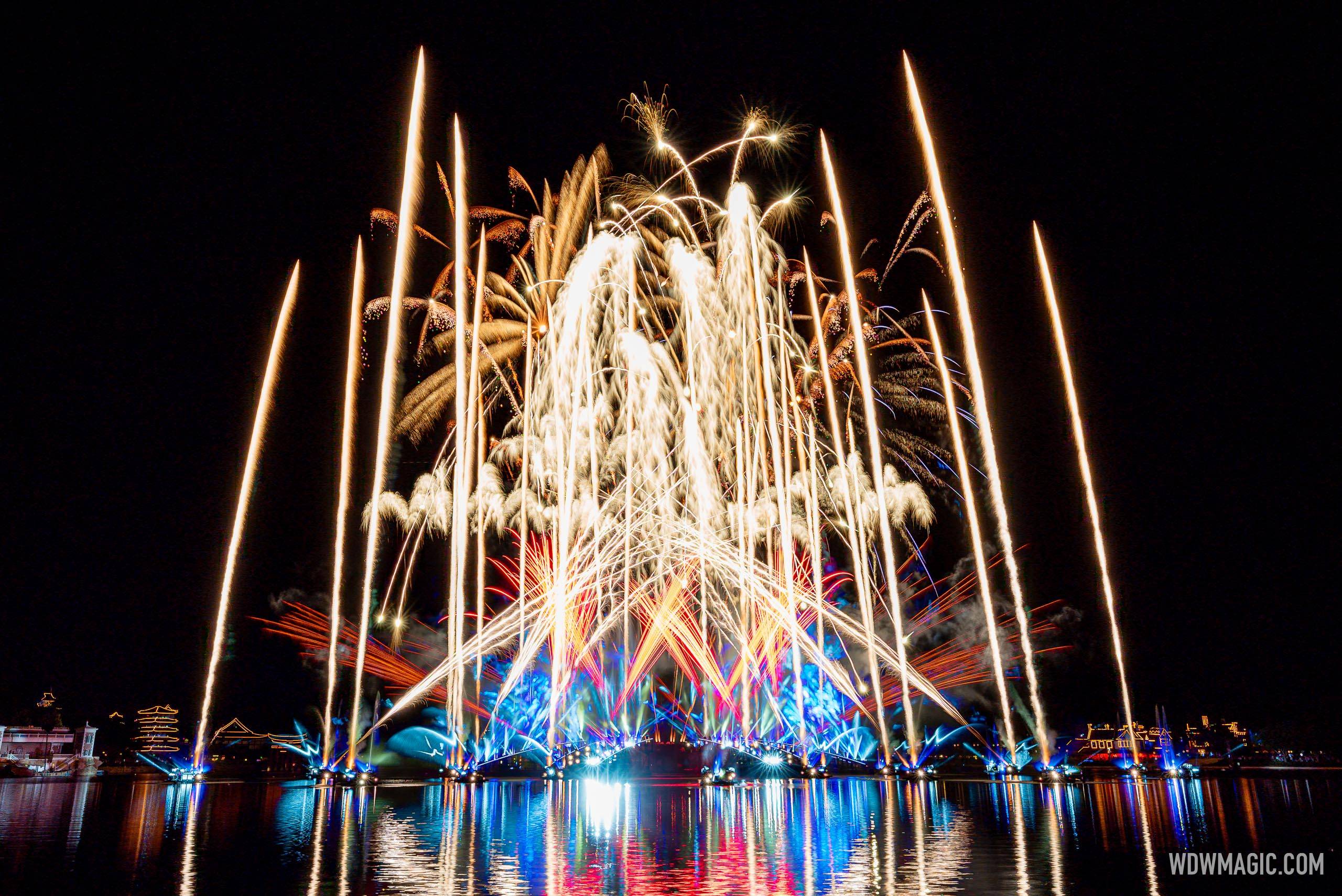 White and gold fireworks and tall streaks of light rise over World Showcase Lagoon at night, with blue and red lights glowing on the water and reflections below