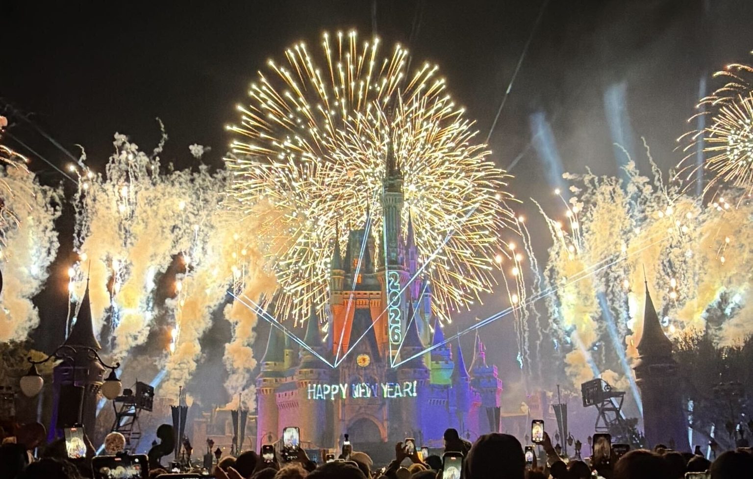 Gold fireworks burst over Cinderella Castle, which has 2026 projected down its spire and HAPPY NEW YEAR! projected across its base, with a crowd holding up phones in the foreground