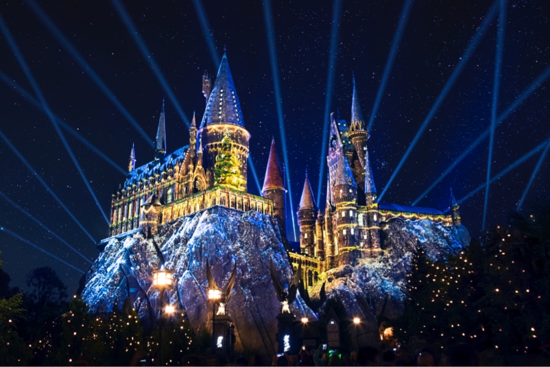 Hogwarts castle at night with festive projections across its walls, a lit Christmas tree on the facade and searchlight beams in the starry sky