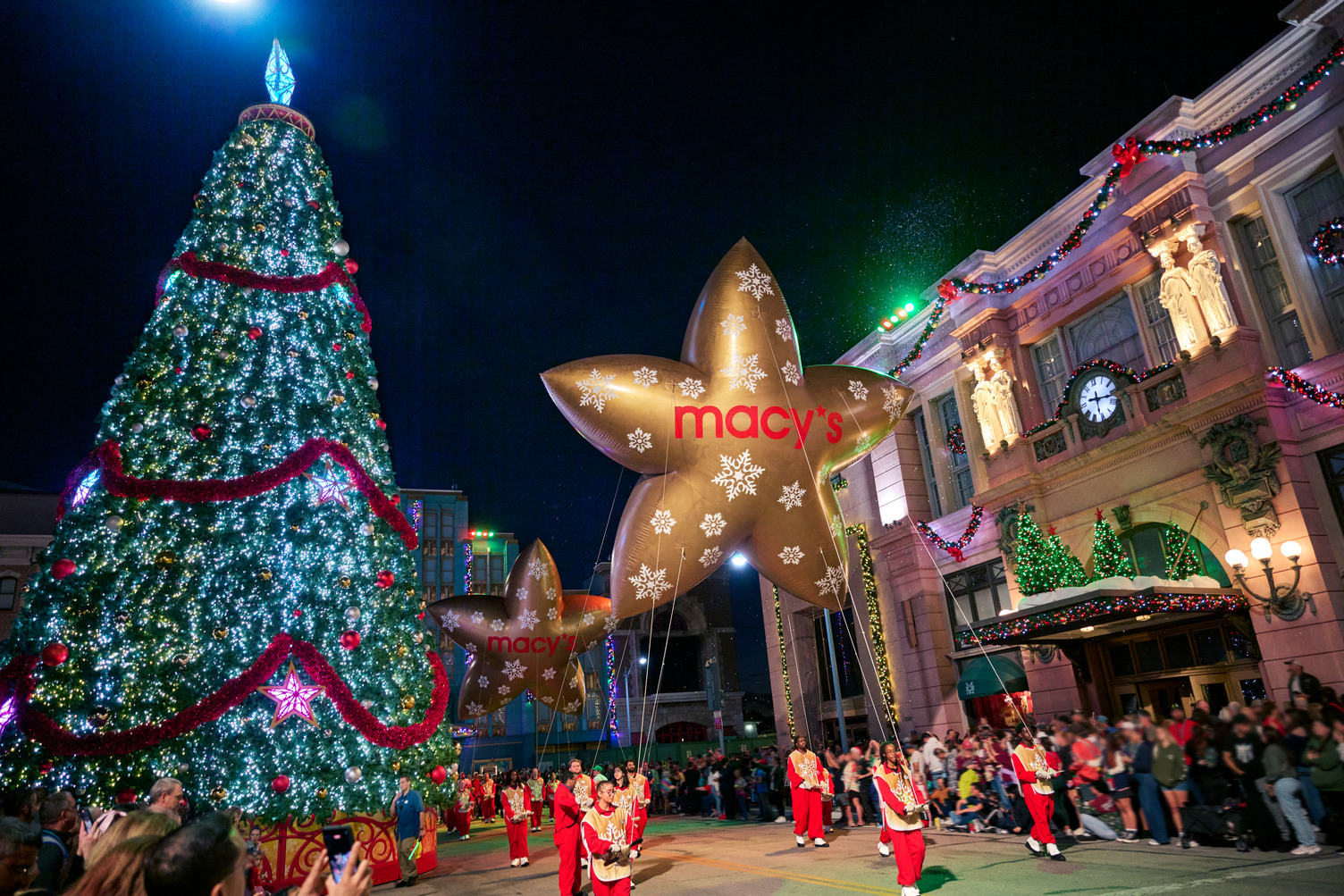 Gold star balloons with the Macy's logo float down a street at night past a giant lit Christmas tree and a garland-trimmed building, with performers in red and white below