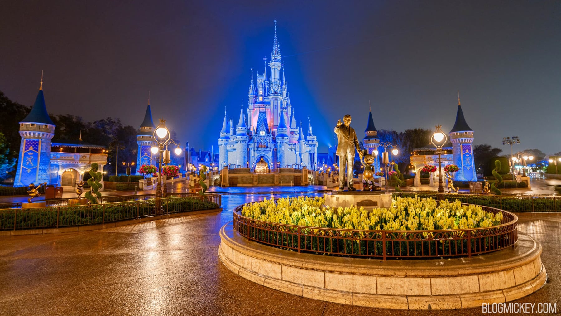 Cinderella Castle lit blue and pink at night behind the Partners statue of Walt Disney and Mickey Mouse, with a bed of yellow flowers in front and wet pavement reflecting the lights