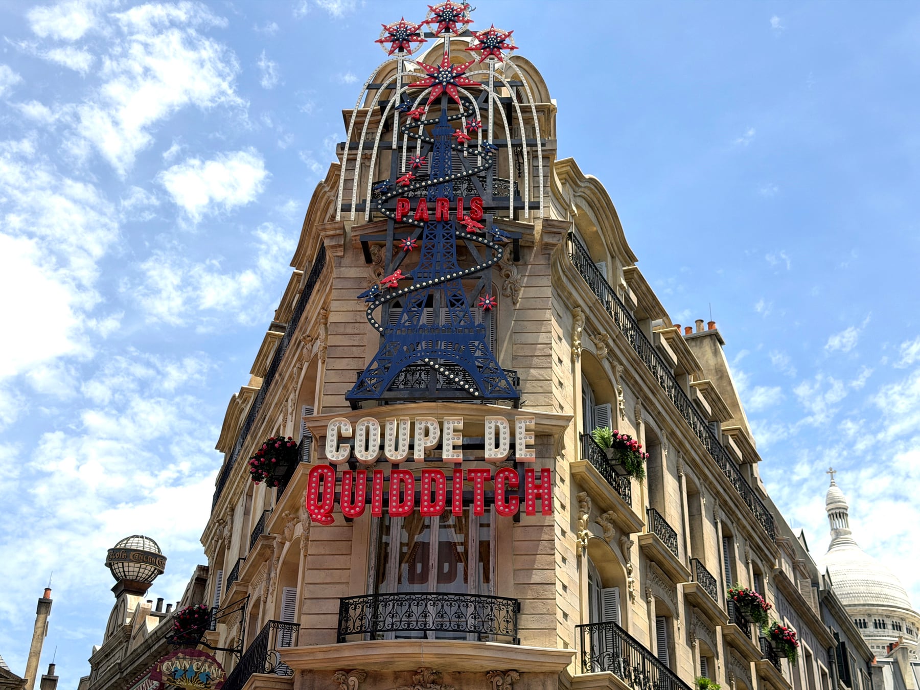 A Parisian-style corner building with a blue Eiffel Tower sculpture on top and a large marquee sign reading Coupe de Quidditch in white and red letters, against a blue sky