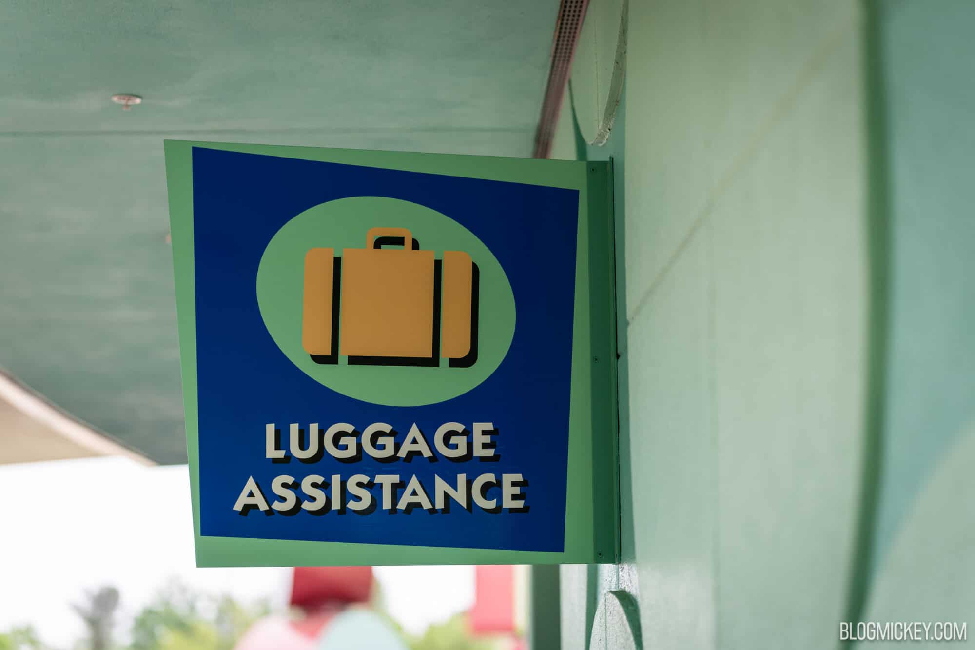 A blue and green Luggage Assistance sign with an orange suitcase icon, mounted on a mint green wall under an overhang
