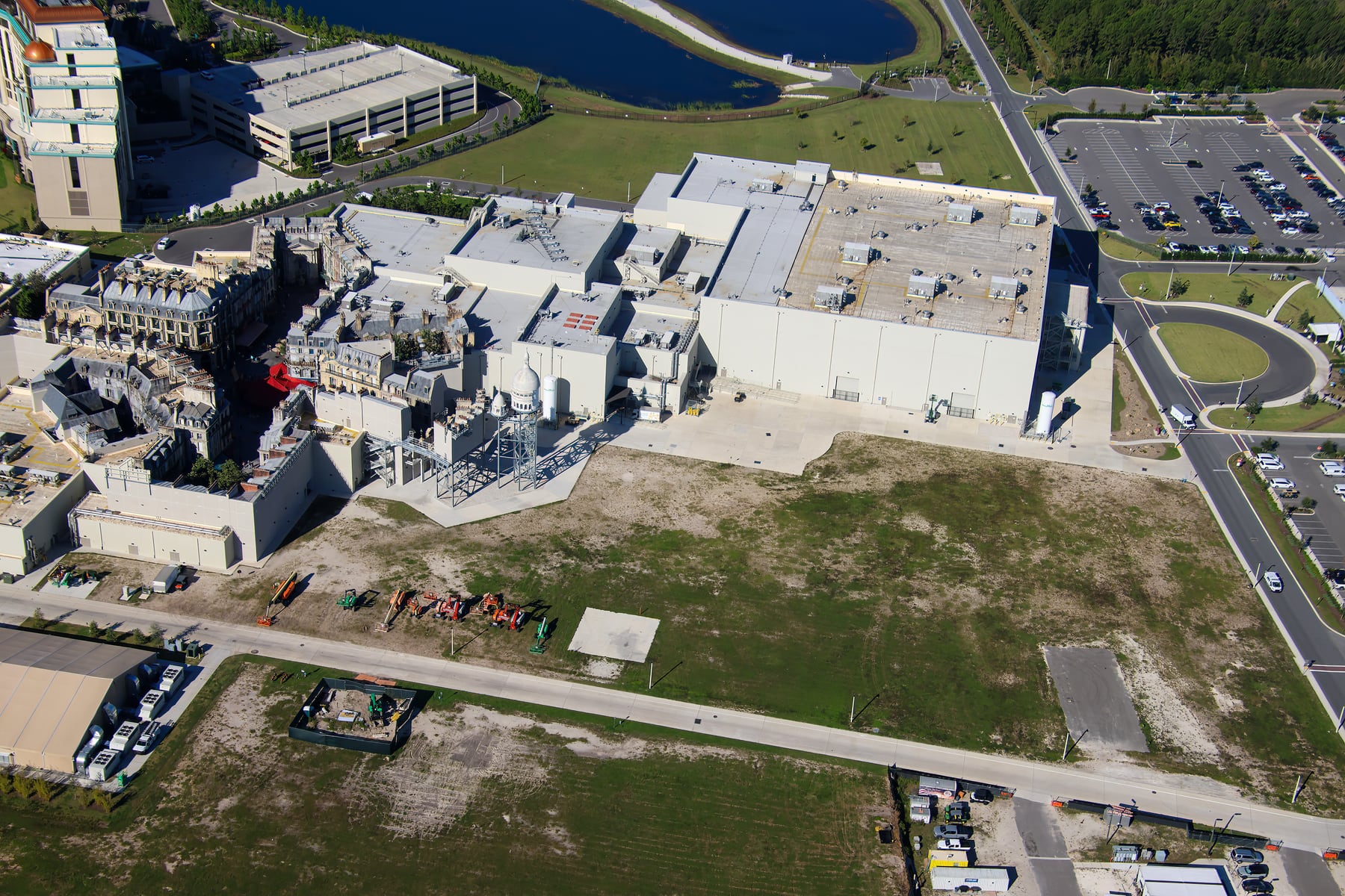 Aerial view of the large white show buildings of the Wizarding World at Epic Universe, with Parisian-style rooftops at left and a wide open grass and dirt lot in the foreground with equipment parked along its edge