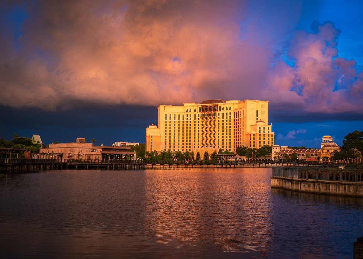 Gran Destino Tower at Coronado Springs glowing gold at sunset under pink and gray clouds, seen across the lake with a Lago sign at left