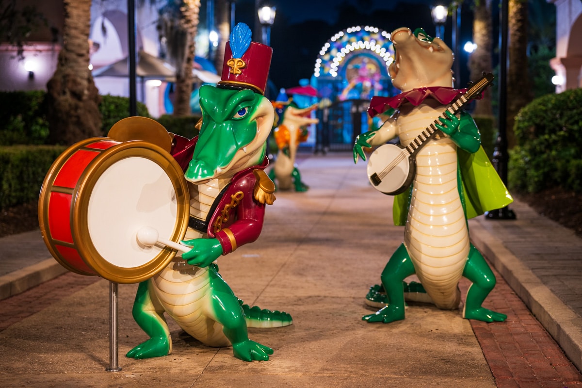 Two alligator band statues at night on a walkway at Port Orleans French Quarter, one playing a bass drum in a red band uniform and plumed hat and one playing a banjo in a green cape, with a lit arch behind them