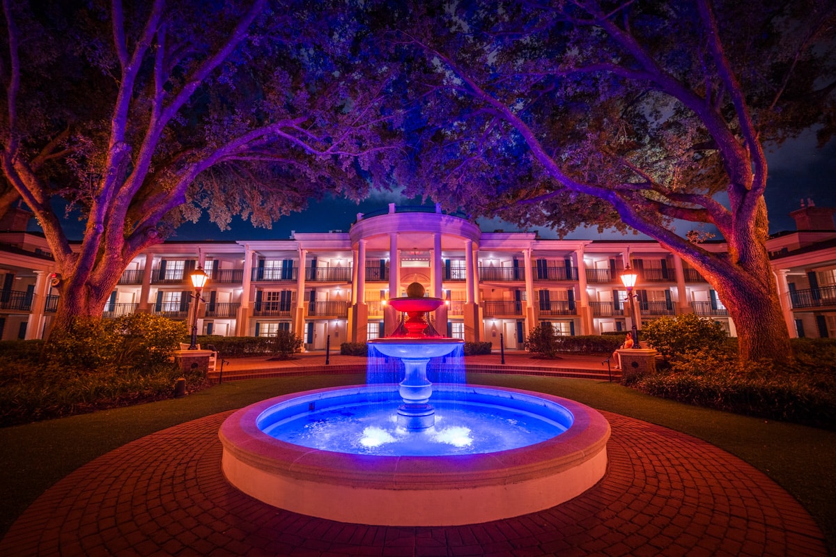 A tiered fountain lit blue in front of a white columned mansion-style building at Port Orleans Riverside at night, framed by large oak trees lit purple