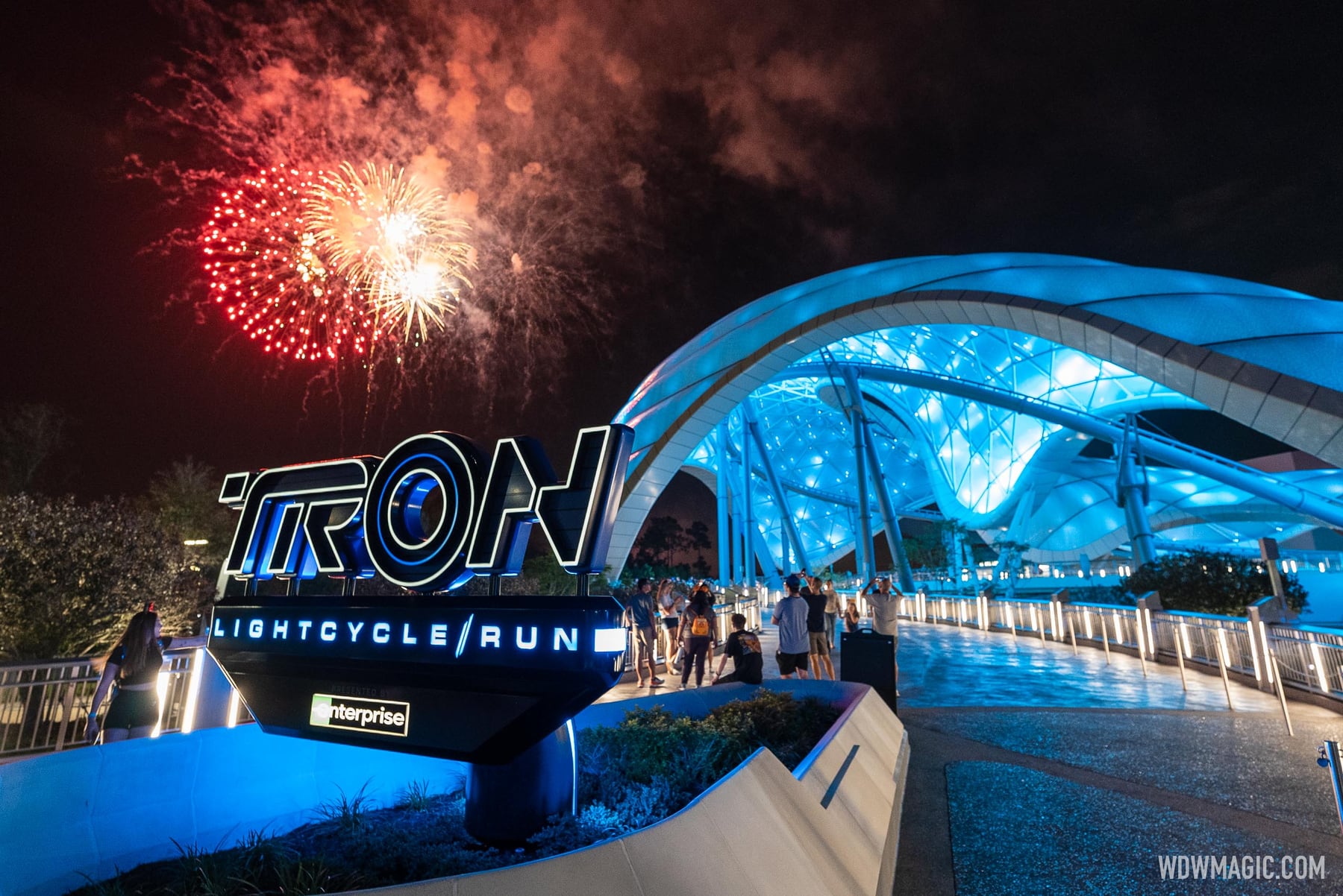 The TRON Lightcycle / Run sign and the glowing blue canopy of the coaster at night, with red and gold fireworks bursting in the sky and guests walking along the path
