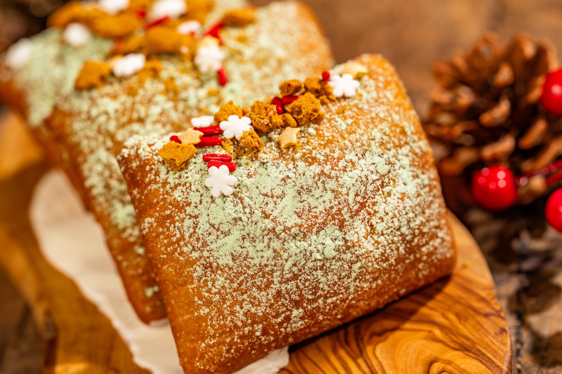 Square beignets dusted with powdered sugar and topped with gingerbread crumbs, red and white sprinkles and snowflake candies, beside a pine cone and red berries